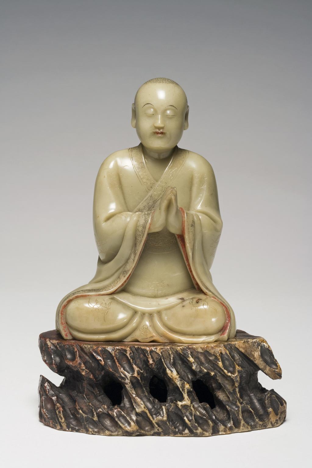 An image of Carving. Soapstone, carved as a seated luohan with his eyes closed and his hands together in a respectful gesture. The collar, sleeves and hem of his robes incised with a design of scrolling lotus on a formalized wave ground. Soapstone, height, including base, 17.5 cm, 1700-1800, Chinese.