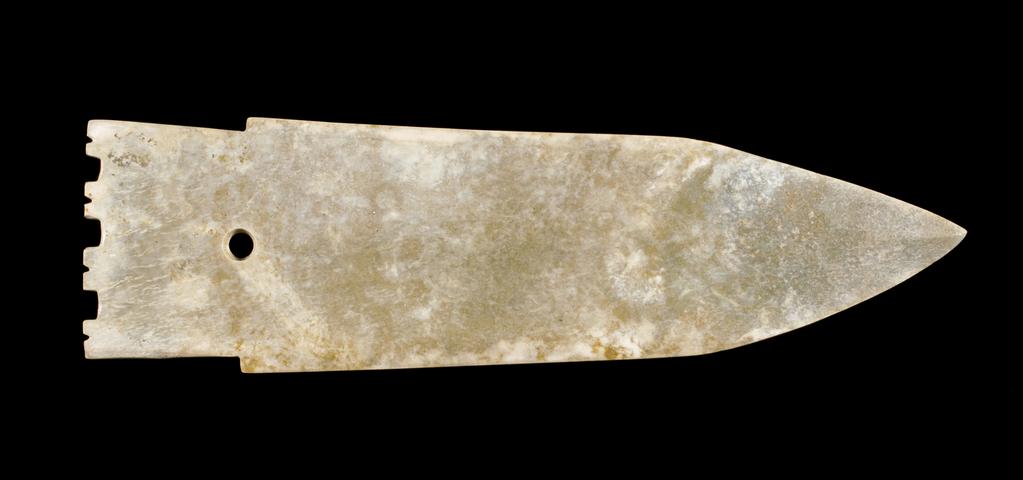 An image of Jade Dagger. Green jade extensively altered to creamy white, carved as a ceremonial blade, in the shape of a bronze halberd, ge. There is a raised central rib and the two edges are bevelled. A perforation is drilled from one side near the tang. Jade/nephrite, length 24.5 cm, width 5.5 cm, 1050-771 B.C. Chinese.