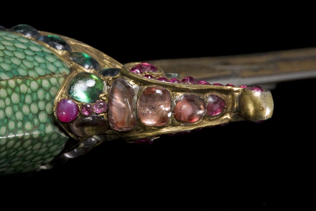 An image of Dagger and Scabbard (khanjar). The blade is straight and double edged, fretted with two slots at either side of a medial serpent, and around a lotus bud. The blade is set with four emeralds and five rubies at either side of the forte, and decorated overall with floral panels and inscriptions in thick gold koftgari. The blade bears the date 'sanat 1121'. The hilt comprises a bulbous grip in shagreen, with a medallion of small diamonds in a gilt setting at the centre reading ‘Sultan Sa’id’. The guard has small forward curved quillons set with various stones and gilded, and a pommel beaked at either side, enamelled in blue, red, pink and yellow in a white ground. The scabbard is of wood covered with shagreen, with large top locket and chape decorated in the same enamel, the chape with a large finial gilt with dark blue and turquoise enamel. Length (whole) 410 mm, width (blade) 270 mm, scabbard 310 mm, weight 280g. Blade 18th Century (1709-10), Turkish hilt & scabbard 19th Century. Turkish, Persian.