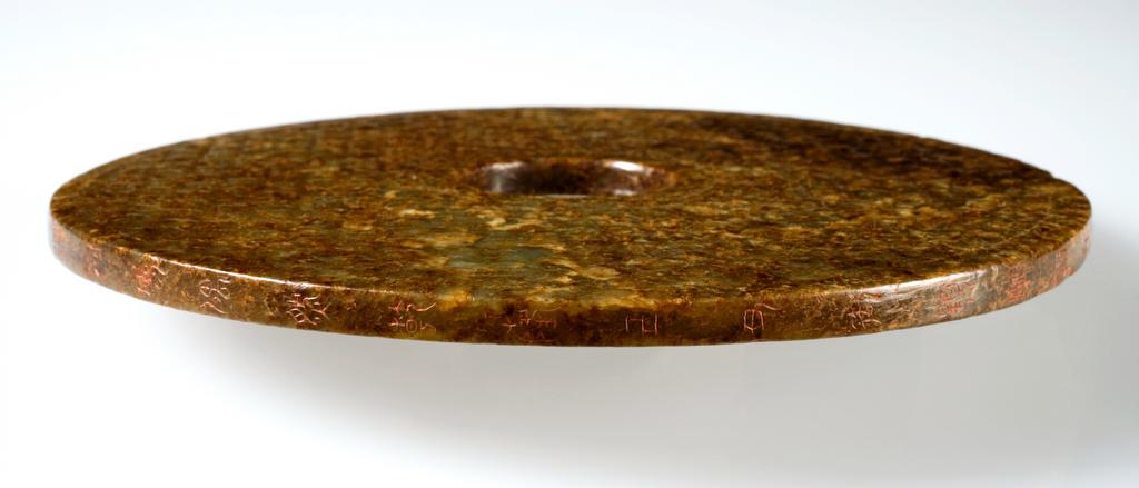 An image of Jade Disc. Grey green jade with brown speckling, decorated with a criss-cross pattern, forming faceted bumps, which are embellished with incised scrolls. The hole in the middle was drilled from one side. The edge of this disc incised with a poem by the Qianlong Emperor, dating to 1769.