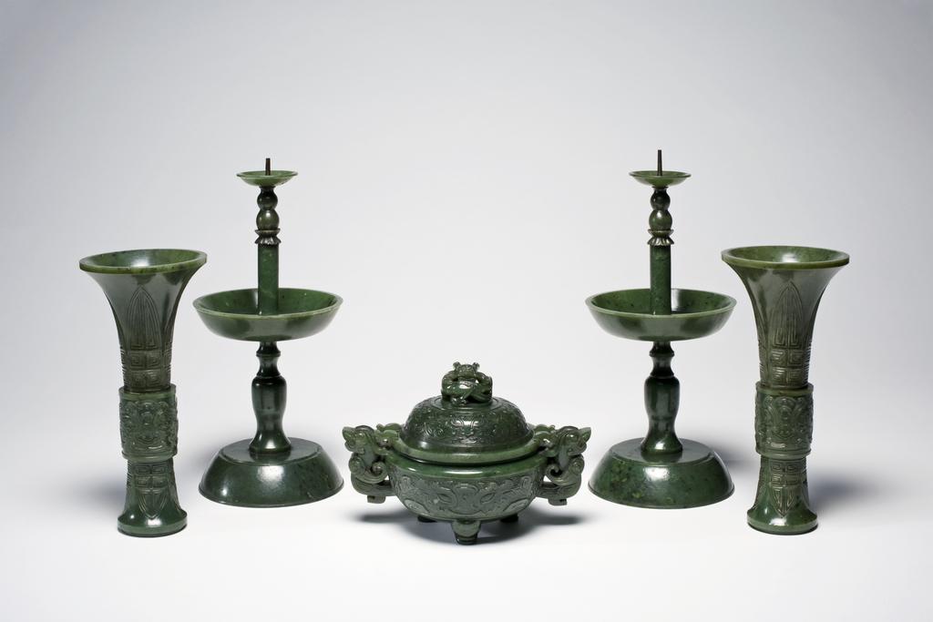 An image of Altar set of five pieces. Dark green with black spots; a Buddhist altar set of five pieces; two pricket candlesticks, two vases, and an incense burner. The two vases and incense burner are carved with ancient jade designs. The set are mounted on an elaborately carved wooden stand. Jasper, 1700-1800. Chinese.