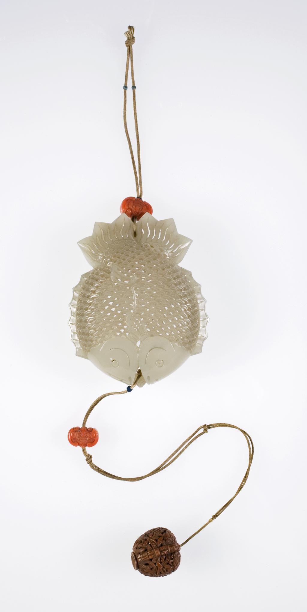 An image of Pomander. Double-fish design. Pale greenish-white jade, carved, pierced and engraved, and made in two halves fitting together as container. Yellow silk cords at each end, on which are threaded bat-shaped coral beads and a carved peach stone. Nephrite, length 7.5 cm, width 6.2 cm, 1700-1800. Chinese.