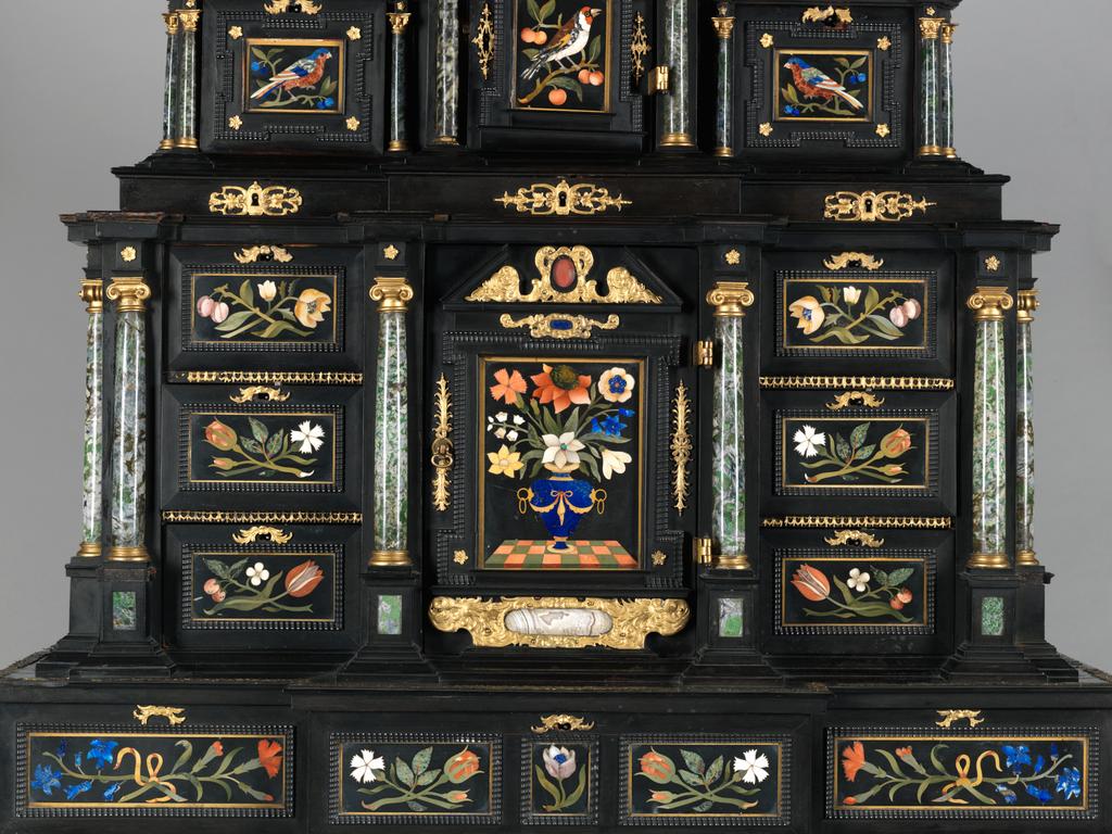 An image of Augsburg 17th century silver-gilt mounted ebony Table Cabinet with Florentine pietre dure plaques from the Grand Ducal workshops. Boscher, Elias (1600-1674). Of architectural form, the stepped superstructure fitted with three frieze drawers above a central cupboard door, architectural pediment and inset with a panel depicting a chaffinch on a fruiting bough, flanked by columns of Smaragdite Gabbro with Corinthian capitals flanked by two deep drawers, inset with panels depicting parrots within fruiting boughs, with three further fitted drawers below, the large central cupboard door inset with a panel of a lapis lazuli flower filled urn issuing stems including a carnation, anemone, daffodil and lilies and enclosing an architectural interior, flanked by further Smaragdite Gabbro Ionic columns and three drawers to each side inset with further symmetrical floral panels including tulips, roses, carnations, lilies and bluebells, on shaped bracket feet, each side inset with large central panels of parrots on fruiting boughs, with four further floral panels above and below, the reverse veneered in kingwood, amaranth and stained walnut in geometric stellar designs. The cabinet containing a complex series of fourty-four secret drawers and compartments to the interior. The cabinet is signed in pencil on the underside 'Elias Boscher' and 'gemacht'. The central cabochon mount stamped with the Augsburg town mark and the silversmiths mark of Johann Spitzmacher (active 1655-1678). The front bears the ebony stamp "EBEN" with the pinecone of Augsburg. Height 85 cm, width 84cm, depth 40cm, circa 1660.