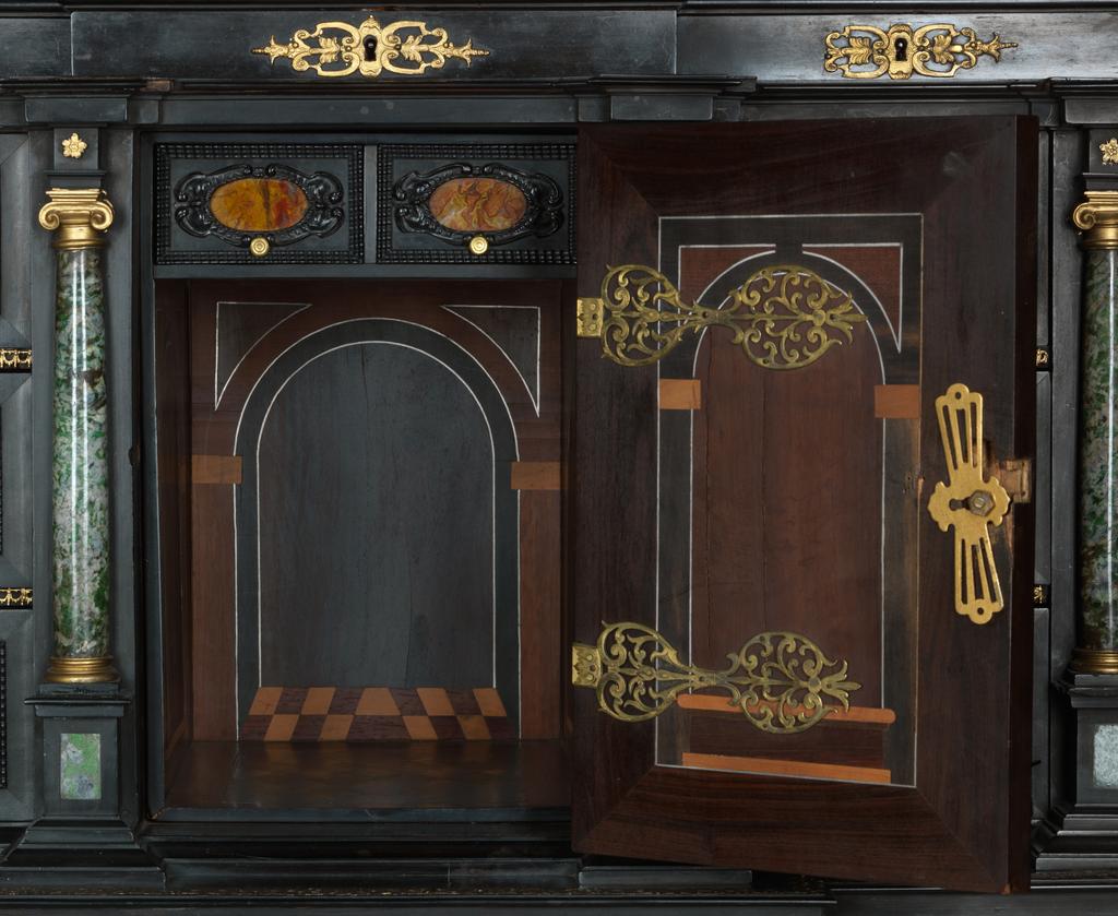 An image of Augsburg 17th century silver-gilt mounted ebony Table Cabinet with Florentine pietre dure plaques from the Grand Ducal workshops. Boscher, Elias (1600-1674). Of architectural form, the stepped superstructure fitted with three frieze drawers above a central cupboard door, architectural pediment and inset with a panel depicting a chaffinch on a fruiting bough, flanked by columns of Smaragdite Gabbro with Corinthian capitals flanked by two deep drawers, inset with panels depicting parrots within fruiting boughs, with three further fitted drawers below, the large central cupboard door inset with a panel of a lapis lazuli flower filled urn issuing stems including a carnation, anemone, daffodil and lilies and enclosing an architectural interior, flanked by further Smaragdite Gabbro Ionic columns and three drawers to each side inset with further symmetrical floral panels including tulips, roses, carnations, lilies and bluebells, on shaped bracket feet, each side inset with large central panels of parrots on fruiting boughs, with four further floral panels above and below, the reverse veneered in kingwood, amaranth and stained walnut in geometric stellar designs. The cabinet containing a complex series of fourty-four secret drawers and compartments to the interior. The cabinet is signed in pencil on the underside 'Elias Boscher' and 'gemacht'. The central cabochon mount stamped with the Augsburg town mark and the silversmiths mark of Johann Spitzmacher (active 1655-1678). The front bears the ebony stamp "EBEN" with the pinecone of Augsburg. Height 85 cm, width 84cm, depth 40cm, circa 1660.