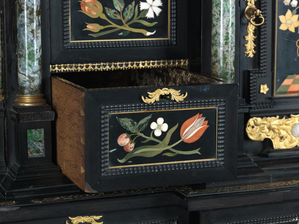 An image of Augsburg 17th century silver-gilt mounted ebony Table Cabinet with Florentine pietre dure plaques from the Grand Ducal workshops. Boscher, Elias (1600-1674). Of architectural form, the stepped superstructure fitted with three frieze drawers above a central cupboard door, architectural pediment and inset with a panel depicting a chaffinch on a fruiting bough, flanked by columns of Smaragdite Gabbro with Corinthian capitals flanked by two deep drawers, inset with panels depicting parrots within fruiting boughs, with three further fitted drawers below, the large central cupboard door inset with a panel of a lapis lazuli flower filled urn issuing stems including a carnation, anemone, daffodil and lilies and enclosing an architectural interior, flanked by further Smaragdite Gabbro Ionic columns and three drawers to each side inset with further symmetrical floral panels including tulips, roses, carnations, lilies and bluebells, on shaped bracket feet, each side inset with large central panels of parrots on fruiting boughs, with four further floral panels above and below, the reverse veneered in kingwood, amaranth and stained walnut in geometric stellar designs. The cabinet containing a complex series of fourty-four secret drawers and compartments to the interior. The cabinet is signed in pencil on the underside 'Elias Boscher' and 'gemacht'. The central cabochon mount stamped with the Augsburg town mark and the silversmiths mark of Johann Spitzmacher (active 1655-1678). The front bears the ebony stamp "EBEN" with the pinecone of Augsburg. Height 85 cm, width 84cm, depth 40cm, circa 1660.