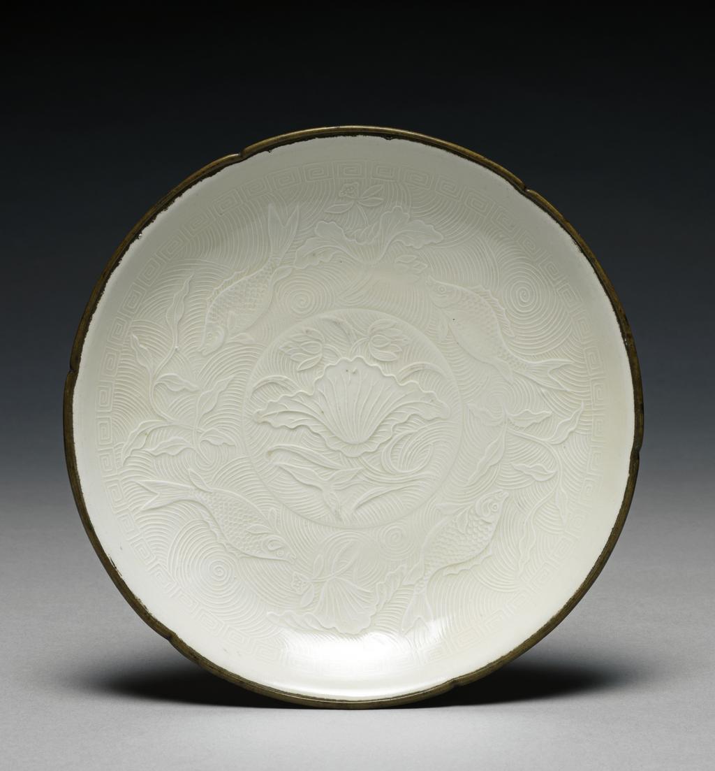An image of Saucer Dish. Ting ware. Dish with a circular foot and bronze rim. Design stamped. Decorated in high relief in the centre with a lotus leaf, flowers and water weeds, on wavy ground. Around this is a band of wave pattern, with four fishes, lotus, and other water plants in high relief. Porcelain, bronze rim, height 3.5 cm, width 19.1 cm. Song Dynasty (960-1279). Chinese.