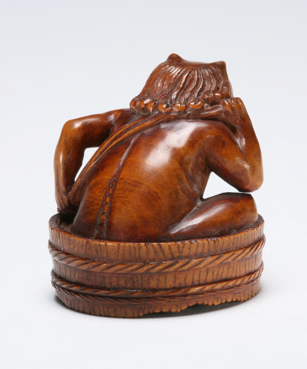 An image of Netsuke: katabori: Oni seated in a bath tub. Hanayama, Edo Period (1603-1868), Japan. An oni sitting in a bath tub drying his back with a towel. Facial features are wide and the mouth is pulled into a grimace. He has two horns on top of his head, a bald central patch and long hair. Himotoshi on the underside. Wood, carved with inalid decoration, height, whole, 3.5 cm, width, whole, 3.5 cm, circa 1800-1868. Edo Period (1615-1868).