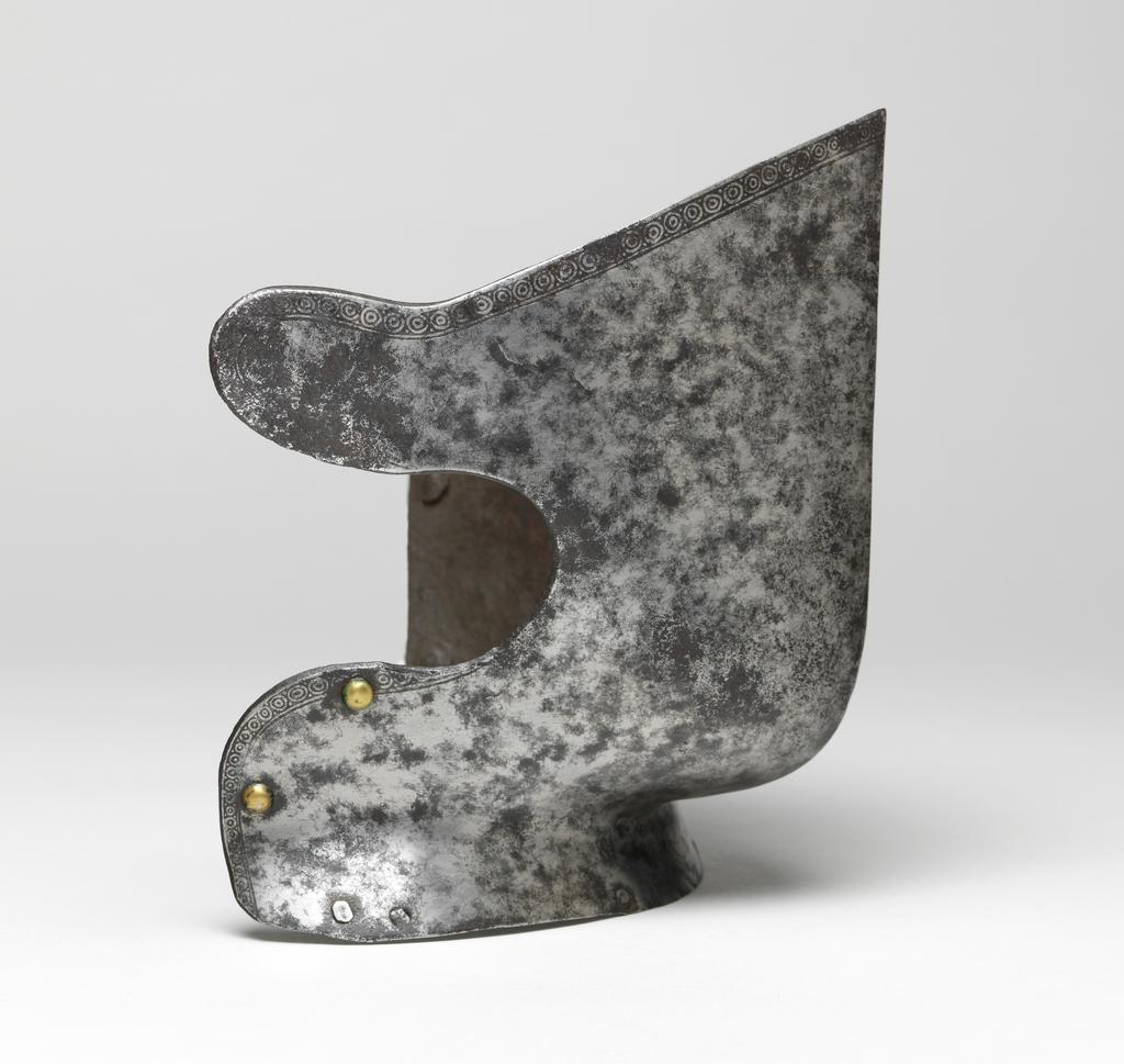 An image of Armour/bevor. Unknown production, South Germany. Reinforcing bevor, for use in the tourney, with etched decoration. Formed of a single, medially-ridged, prow-shaped plate covering the front and sides of the face from the level of the eyes to the top of the neck, and strongly shaped to the chin. Its straight upper edge, forming the lower edge of a vision-slit, slopes upwards at its outer ends to strongly rounded corners. Its sides are nearly vertical. The right side has a large cut-out that may perhaps have been enlarged during the working life of the piece. Located at the right of the neck and at the lower edge of the cut-out are brass-capped, round-headed rivets. Similar rivets with circular, internal washers are located at the left of the neck and half way up the left side. The lower edge of the bevor is flanged outwards to receive gorget-plates. These would have been attached by internal leathers. The outer leathers were secured by pairs of externally-flush rivets, and the inner ones by single externally-flush rivets, all of which survive. The outer ends of the flange are cut off obliquely and slightly rounded. The edges of the bevor, except at the neck and the inner end of the cut-out, are etched with narrow bands of small concentric circles between pairs of pellets, all on a blackened ground. The bevor is bright with heavy, mottled patination and some severe patches of pitting overall. It is rusted through down the centre of the front of the chin. Steel, hammered, shaped, riveted, with etched decoration on a blackened ground. Height, whole, 19.7 cm, maximum, width, whole, 23.2 cm, maximum, depth, whole, 11.6 cm, maximum, weight, whole, 0.66 kg, circa 1550-1560. From the armoury of the Princes Radziwill, Castle of Niescwiez, Poland.