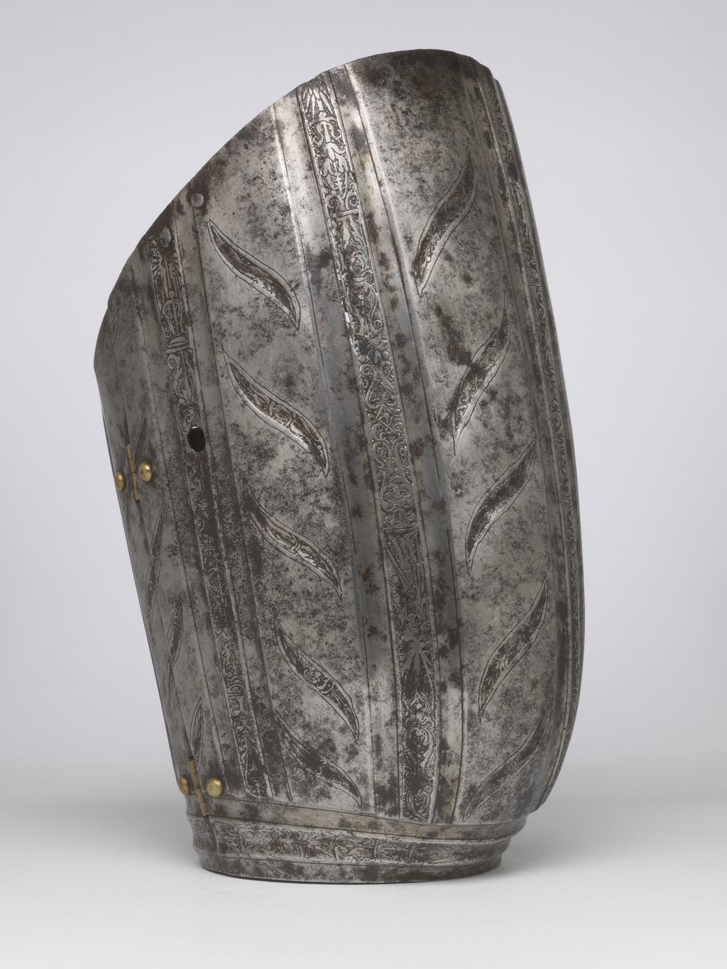 An image of Armour. Couter and lower cannon of a vambrace for the right arm, for parade use, with 'puffed-and-slashed' decoration in imitation of civilian costume. Production Place: South Germany. Helmschmied, Koloman (German, 1470/71-1532). The couter is formed of a single, large, shell-like plate, open at the rear and strongly shaped to the point of the elbow. Its outer edge is nearly straight. Its upper and lower edges are convex and converge at the inside of the elbow in a strong, medial pucker. The upper and lower edges have inward turns, roped with pairs of incised lines. The turns are each accompanied by a recessed border which is itself accompanied on the front by a flute separated from the border by a raised rib emphasised by a pair of incised lines. The boss over the elbow is enclosed by two groups of three flutes, each emphasised by pairs of incised lines, that converge to the inside of the elbow. Two vertical groups of such flutes decorate the front of the couter both above and below the boss, while three more decorate the boss itself. The broad bands intervening between these groups of flutes are decorated with slender, diagonal, slightly recurved, recessed 'slashes' in imitation of those found on contemporary civilian costume. The front and rear of the central boss are each fitted with a pair of horizontally-aligned rivets for the attachment of a missing pair of vertical internal leathers that formerly connected the couter to the upper and lower cannons of the vambrace. The outer end of the couter is filled with a pair of widely-spaced, externally-flush rivets for the attachment of a missing bifurcated strap that terminated in a buckle to receive the missing strap that was formerly retained internally by a brass-capped, round-headed rivet and octagonal internal washer located just to the inside of the medial pucker, and passed around the rear of the elbow. The lower cannon is of tubular form, cut away in a concave curve towards the inside of the elbow, and constri