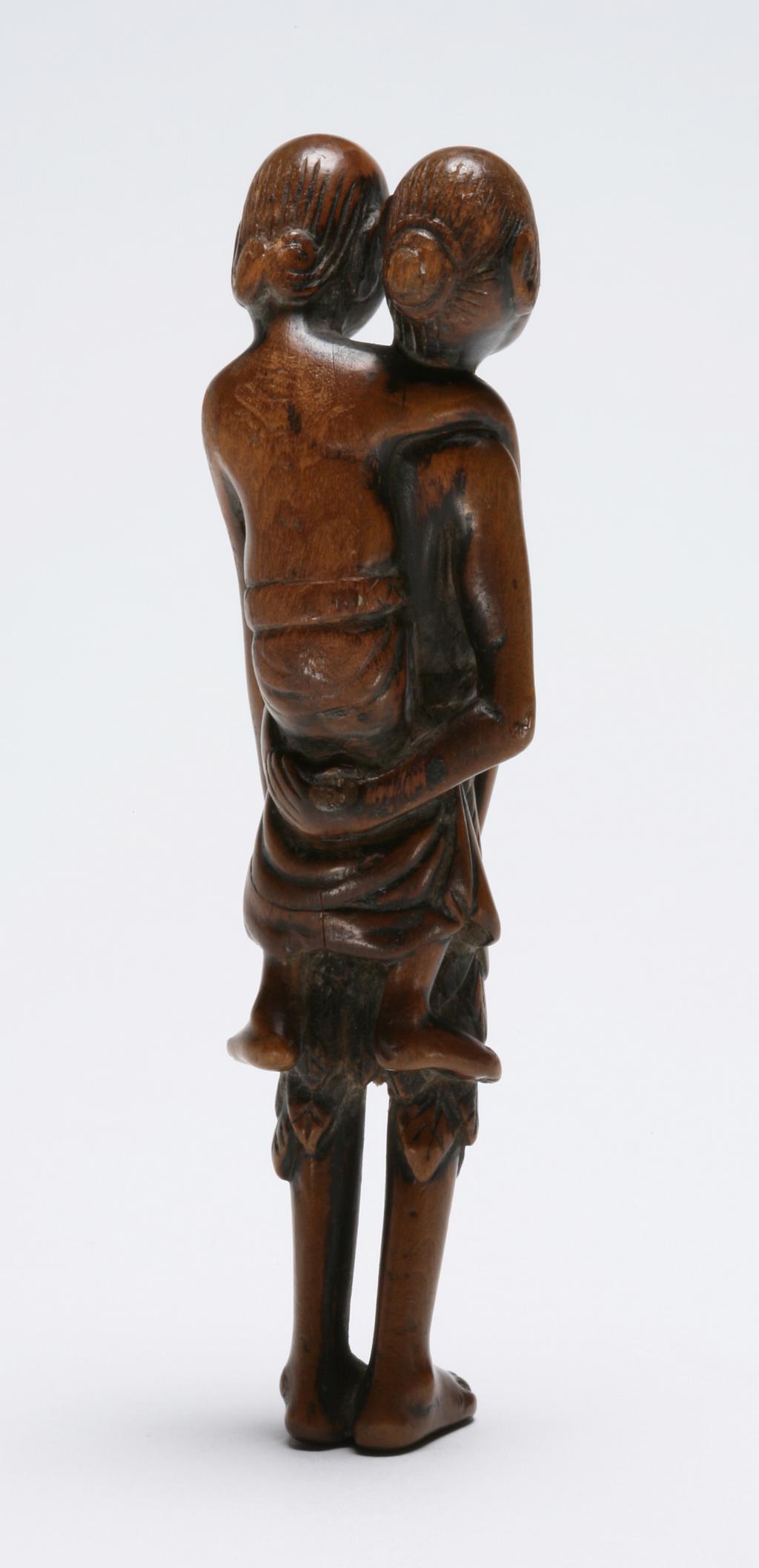 An image of Netsuke: sashi: Ashinaga and Tenaga. Unknown maker, Japan. Ashinaga and Tenaga, the latter seated on his friend's back and reaching down with his elongated arms to hold up Ashinaga's leaf skirt. Ashinaga's hands are clasped behind his back holding up Tenaga. Their hair is scraped into a bun. Natural himotoshi formed by arms and legs. Boxwood, carved and stained, height, whole, 9.9 cm, width, whole, 2.5 cm, circa 1800-1868. Edo Period (1615-1868).