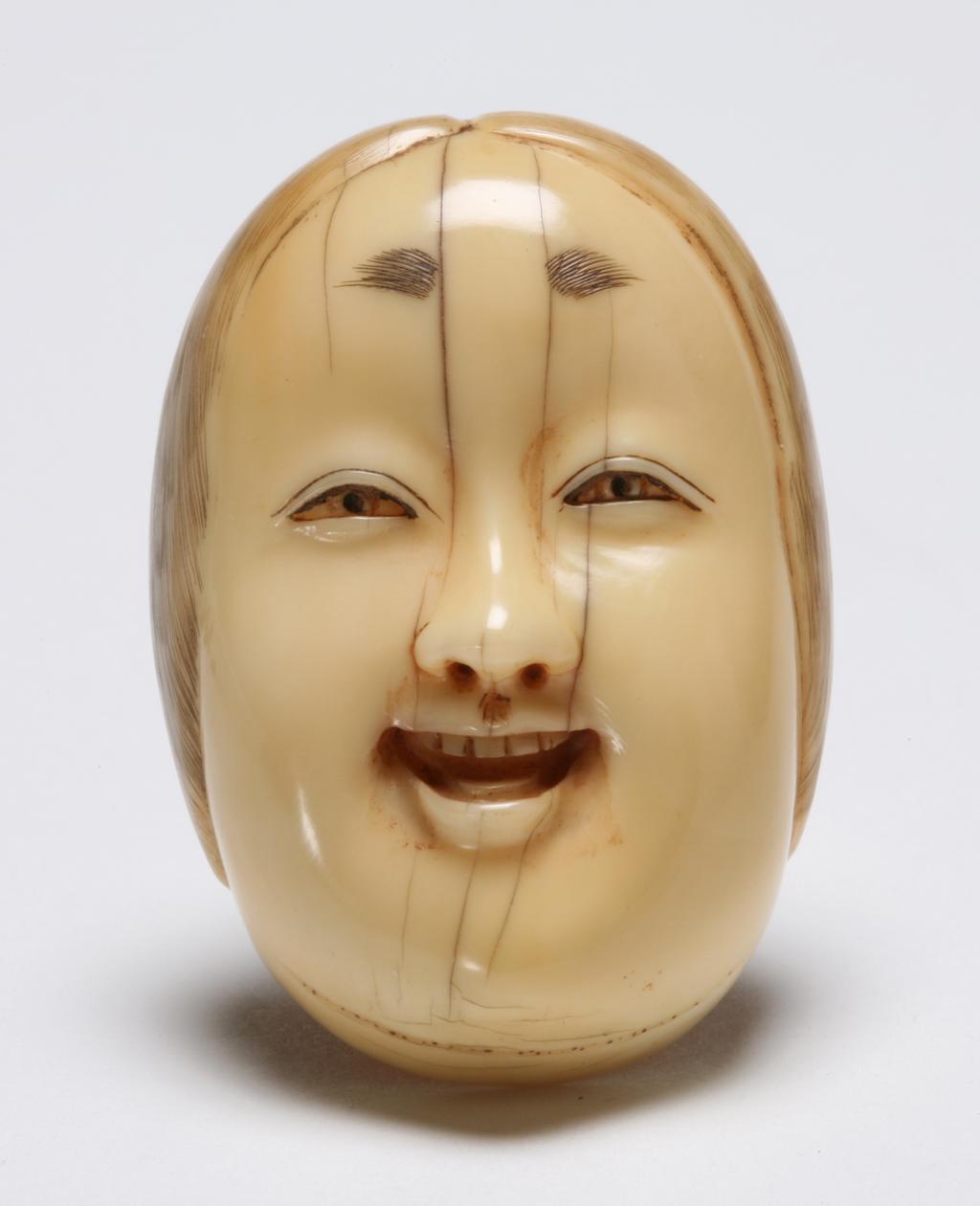 An image of Netsuke. A mask of Okame. Unknown maker, Japan. Ivory, lightly inked hair work and eyebrow, 1870-1930.