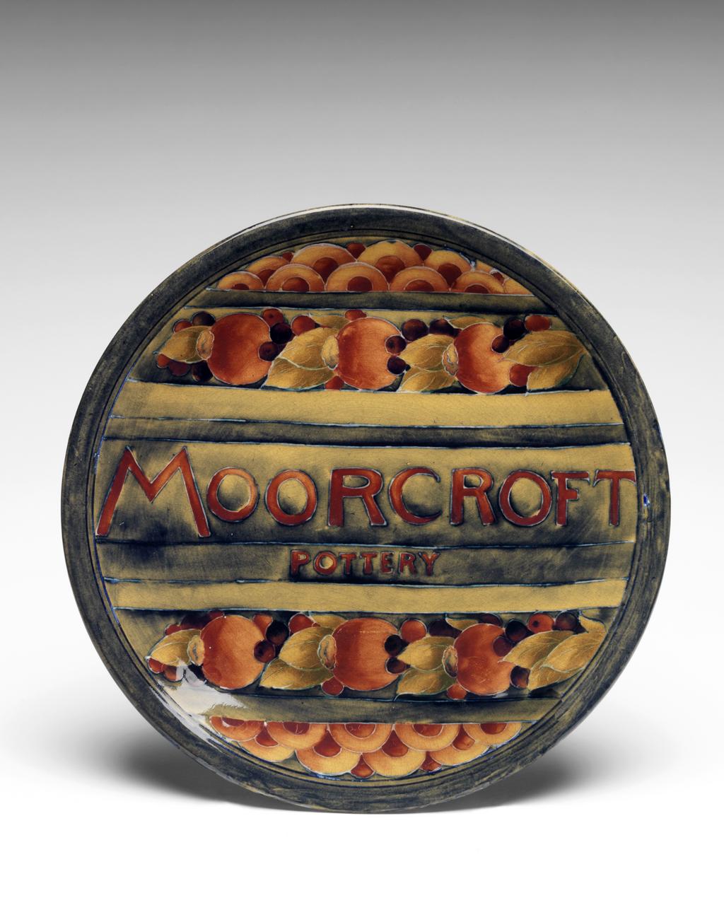 An image of Exhibition Plate. Moorcroft, William (British, 1872-1945). Moorcroft Pottery, Staffordshire, Cobridge. The front is decorated with the words ‘MOORCROFT/POTTERY’ between horizontal bands of pomegranates, berries and leaves, and scale pattern. Cream earthenware plate with slip trailed design, painted underglaze in dull shades of ochre, red, dark red, and blue-black, with 'Pomegranate' pattern, press-moulded, height 3 cm, diameter 26 cm, circa 1913-circa 1920 (probably c.1916). Art Pottery.