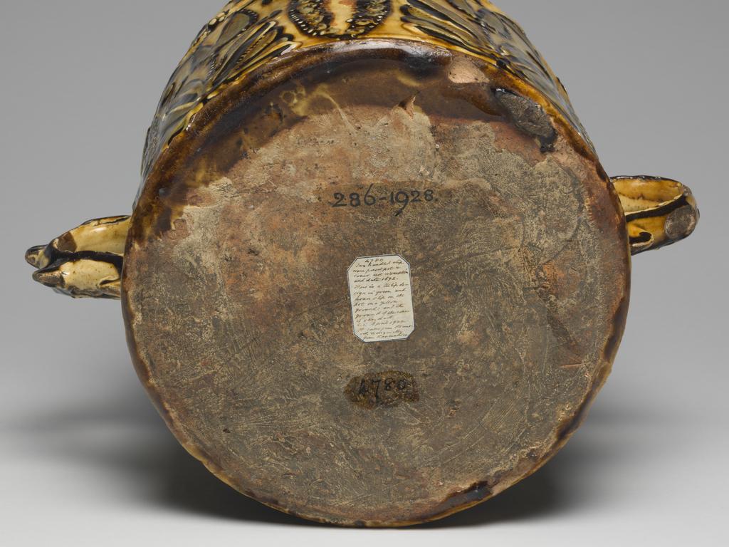 An image of Posset Pot/Two-handled pot and cover. Staffordshire Pottery, unknown maker. Red earthenware, thrown, coated inside and out with white slip, and slip-trailed in dark red, olive-green, and white slips under yellowish lead glaze. Cylindrical with two S-shaped handles. Decorated round the top with a slip-trailed inscription, 'THE BEST IS NOT TOO GOOD FOR YOV 1692'. Below this, on each side, is a pair of stylized tulips separated by vesica-motifs, the latter made up of studs of slip with star impressions of a cut stick. The lid is left red without a coating of white, and is decorated with quatrefoils in white ouline filled in with olive-green slip showing the impression of a textile mesh. 1692. William III and Mary II. No dimensions recorded. Production Note: The cover is almost certainly a 'marriage'.