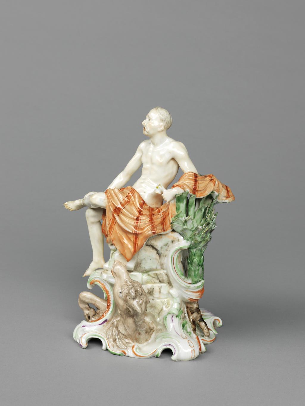 An image of Figure. Asia from a set of Continents. Nymphenburg Porcelain Factory, Germany, Bavaria. Auliczek the Elder, Dominikus Jacob, modeller (Bohemian, 1734-1804). The square base has foliated corners and scrolled sides, and rises up into a tall pedestal of masonry and rocks painted in shades of pale green and grey. Attached to it on the viewer’s right is a C-scroll, and green stylized palm with three visible brown roots. A pale brown camel’s head tilted upwards projects from the base at the front, and one of its foreleg projects from a C-scroll to the left of it. Asia is represented by a thin and palid elderly man with short grey hair and moustache, and black eyes. He sits on the pedestal with his left leg over the viewer’s left edge, and his right crossed over it. His right hand rests on the calf of that leg, and he leans back on his left which is supported by the top of the palm tree. He is nude apart from a drape which is striped in pale and darker reddish-brown. Hard-paste porcelain, press-moulded, and painted overglaze in shades of green, flesh, pale brownish-red, brownish-red, purple, pale and dark brown, grey, and a little black. The underside is hollow and glazed apart from the outer edge, and there is a circular ventilation hold under the camel’s head. Height, whole, 18.4 cm, width, whole, 13.5 cm, circa 1765. Rococo. Production Note: Auliczek succeeded Bustelli as chief modeller at Nymphenburg in 1763, and remained there until 1797.
