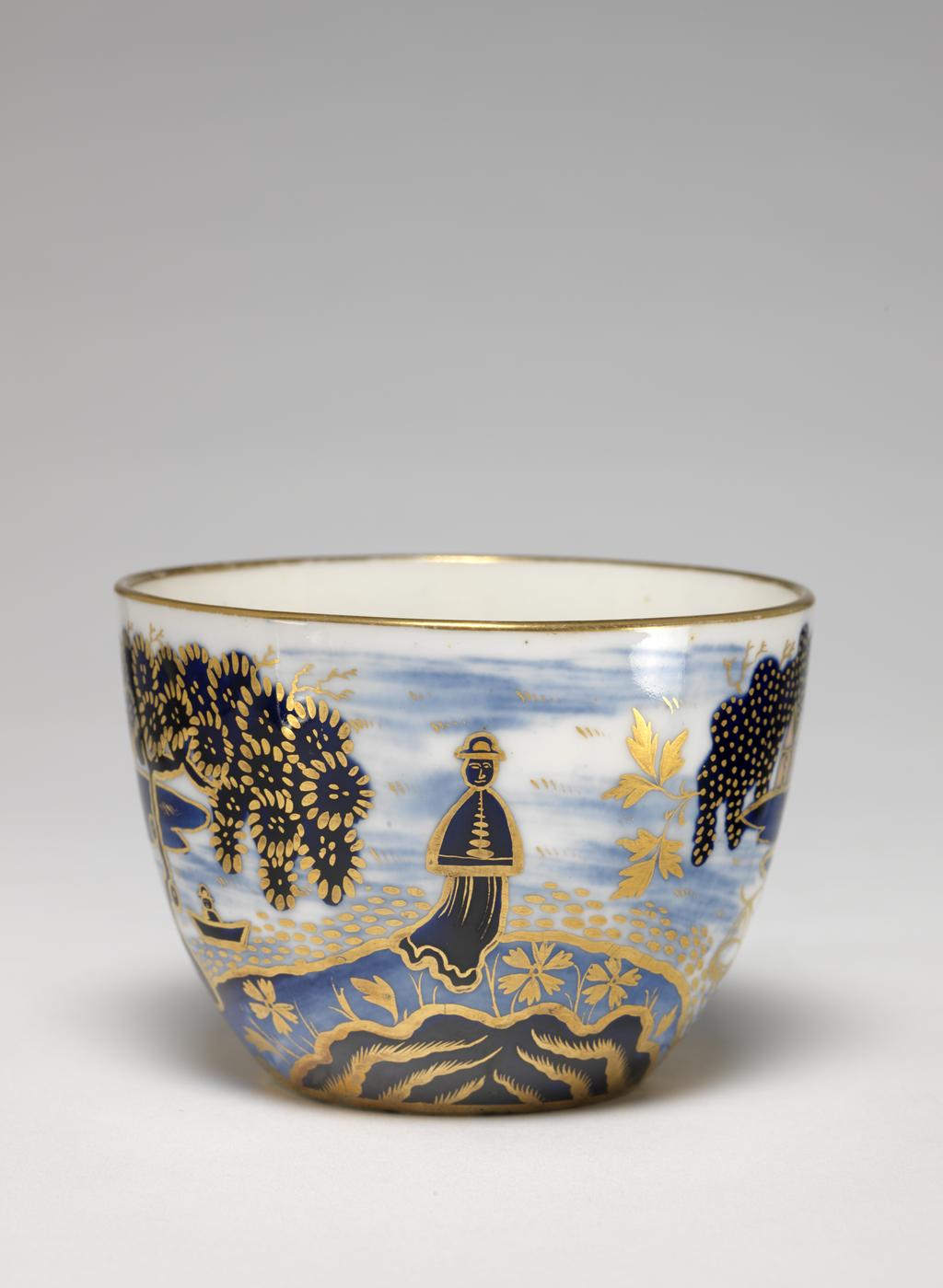 An image of Cup and Saucer/Teacup. New Hall Porcelain Factory, Staffordshire. The cup is of Bute shape with oval ring handle. The saucer is circular with curved sides, standing on a footring. The cup is decorated on the exterior with an Oriental landscape with buildings on the left and right, a man standing fishing on a bridge, a man in a boat, and a larger man in the middle. Inside below the rim there is a continuous horizontal spray of leaves in gold. There is a gold band round the rim, and six stylized leaves down the back of the handle. The interior of the saucer is decorated to match the exterior of the cup and has a gold band round the rim. Pattern 1163. Bone china, painted underglaze in blue, overglaze in apricot-pink enamel, and gilded. Height, cup, 6 cm, width, cup, 10.5 cm, diameter, cup, 8.5 cm, height, saucer, 3.5 cm, diameter, saucer, 14.5 cm, circa 1815-1820. Imari style.