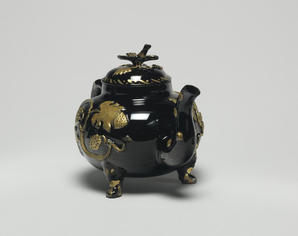 An image of Teapot and cover. Unidentified Staffordshire Factory. Dark red earthenware, the body thrown, with applied moulded handle, spout, feet, and finial, and applied moulded sprigs, the whole covered with black lead-glaze, and partly gilded. The pot has a depressed globular body supported on three lion's mask and paw feet, a crabstock handle and spout, and a domed cover with a bird finial. Both sides of the body are decorated with a coiling vine-stem with bunches of grapes and foliage in relief, which have been oil gilded. Rococo. No dimensions on Adlib (07/2014).