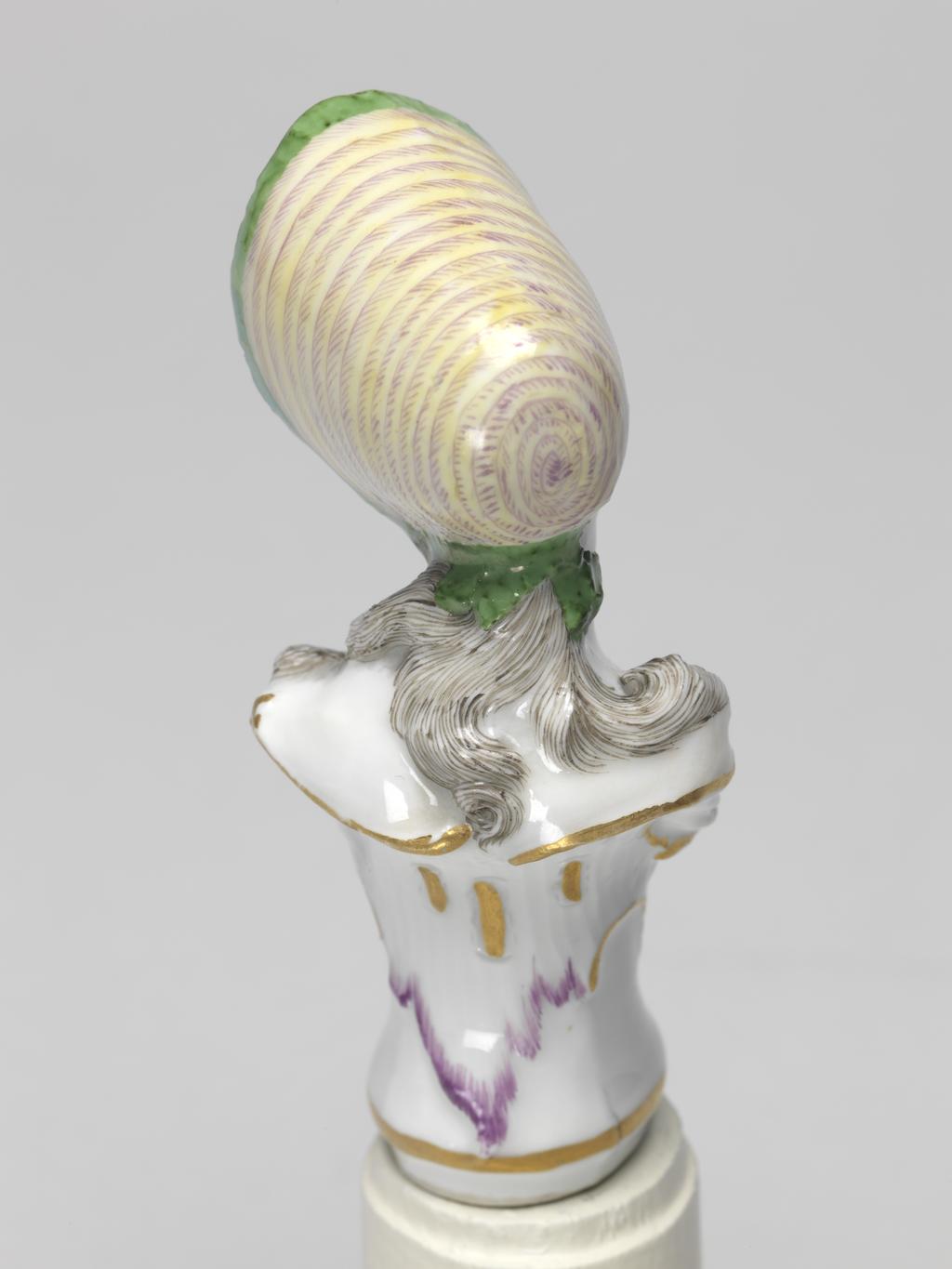 An image of Cane Handle. Nymphenburg Porcelain Factory, Bavaria. Bustelli, Franz Anton, modeller (Swiss, 1723-1763). In the form of a bust of a young woman with an integral, waisted sockle, with lightly fluted sides, and scrolls below her bust and round the back of her shoulders. The young woman's head is turned and tilted slightly to her left. She has long curling black hair which hangs down over her shouilders at the back and sides. She wears a yellow and purple striped bonnet with a green underside to the brim, and green ribbons tied under her chin, and a white bodice or chemise. The sockle is decorated with an inverted leaf-shaped outline in blue on the front, and a purple one on the back, The scrolls round its upper edge are gilded, and there is a horizontal gold band just above the lower edge. Hard-paste porcelain, press-moulded, glazed, and painted in blue, green, yellow, flesh, a little red, purple, and black enamels, and gilded; the interior is hollow and unglazed. Height, whole, 8 cm, width, shoulders, 3.5 cm, circal 1769-1760. Rococo.