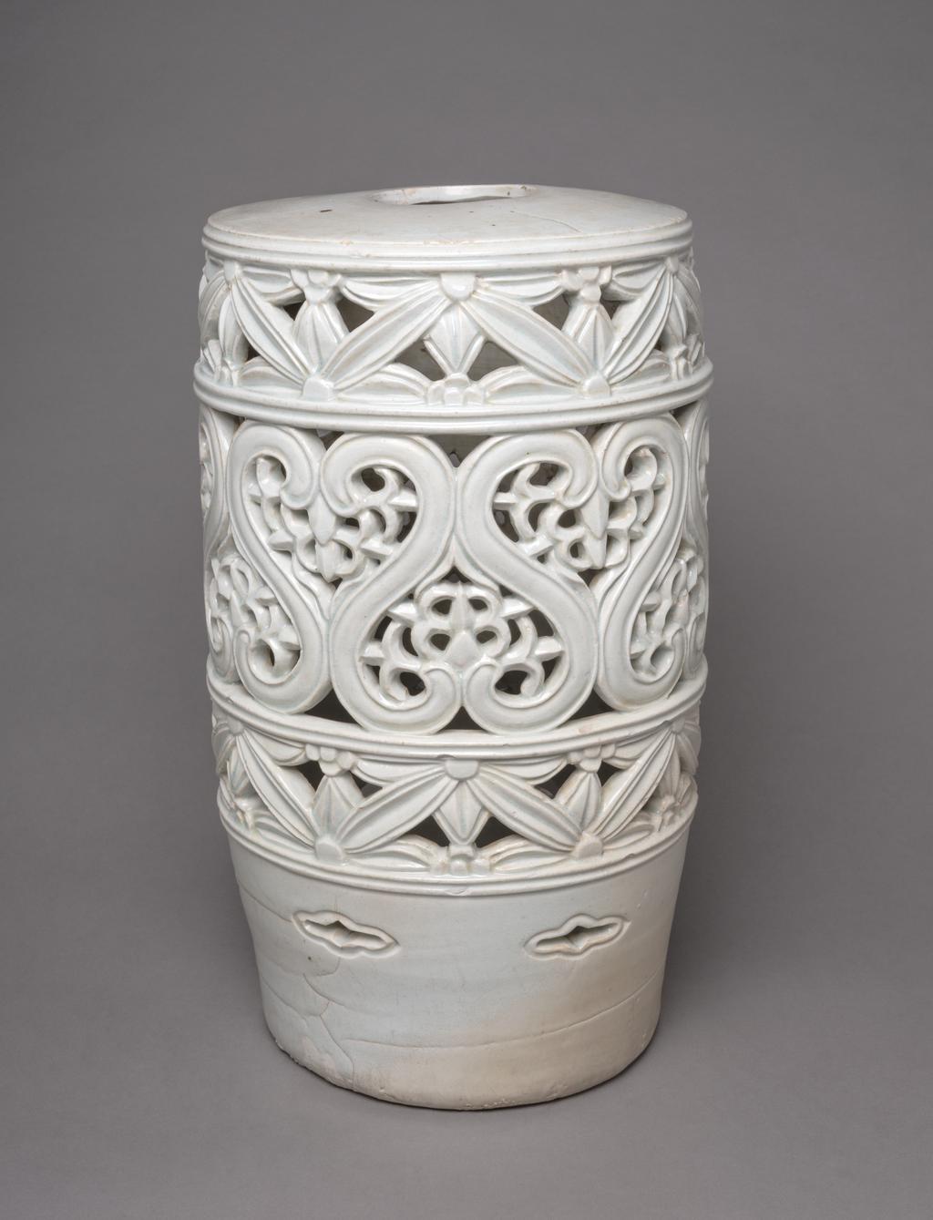 An image of Stand with floral designs. Unknown pottery, Korea. This large stand is cylindrical and has a hole in the top. The sides are divided into three horizontal zones and decorated with two bands of pierced floral designs reminiscent of Korean latticework, around a wider central pierced band with unusual S-shaped motifs enclosing leaves; above the base are small eye-shaped (ansang) openings. The glaze has a pale bluish tone. The stand has no base and the footring shows remains of coarse sand from the firing support. Porcelain, thrown, carved, pierced and glazed, height, whole, 48.7 cm, diameter, top, 26.3 cm, diameter, foot, 23.5 cm, circa 1800-1900. Choson Dynasty (1392-1910). Production Note: This piece was probably used as a stand for a flowerpot.