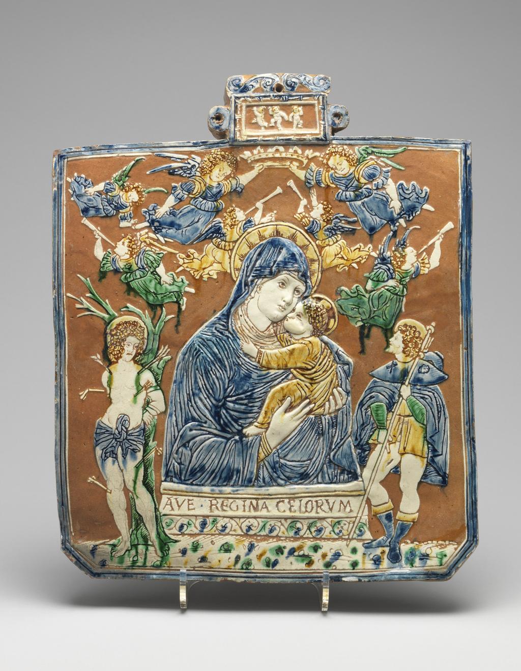 An image of Panel. The Virgin with Saints Roch and Sebastian. Unknown, perhaps The Veneto. Almost square with cut lower corners and a rectangular, scrolled protrusion at the top, with lateral rings and a hole for suspension. A half-length figure of the Virgin holding the Christ Child in her arms, with below, a pedestal inscribed 'AVE.REGINA CELORVM' (Hail Queen of the Heavens), and above, seven angels, of whom two hold a crown over her head and four blow trumpets. St Roch stands on the right hand and St Sebastian on the left. The small panel at the top contains three dancing putti. Pale red earthenware, moulded in very low relief in the middle, and coated with white slip which has been removed from the background. Incised decoration coloured blue, green and dark yellow under lead glaze. Height, whole, 33.7 cm, width, whole, 27.8 cm, depth, whole, 1.7 cm, circa 1500-1510. Renaissance.