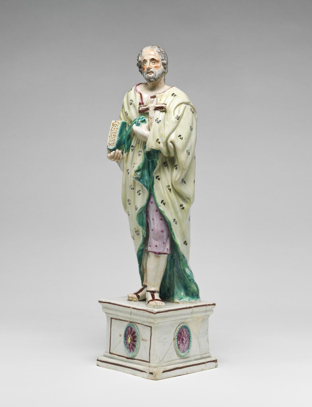 An image of Figure of St. Peter. Standing, wearing a flowered robe, holding a book and a cross, on a high pedestal with his name on the front. Lead-glazed earthenware. England, Staffordshire.