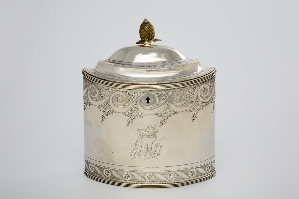 An image of Tea Caddy. Bateman, Peter. Bateman, Ann. Bateman, William. Production Place: London. Bright cut borders and engraved armorials. Silver; the oval body with two compartments, beneath a stepped and slightly domed cover surmounted by a stained bone pineapple finial on a silver cast leaf-shaped calyx. The lid is secured by a lock at the centre front, opposite a concealed hinge. The top and lower edge of the caddy have applied bands of reeding. The sides are decorated with a lower border of alternating roundels and leaves reserved in a striated band with narrow bands of engine turning above and below; the upper border is of bright-cut paterae, Vitruvian scrolls and foliage. The lid has borders of machine engraved decoration. Below the lock is a crest of a boar's head over the initials 'JML'. On the back is engraved a coat of arms, sable goutté argent, a canton ermine. The silver was flattened by rolling. The body was then cut from the sheet and seamed at the side. The cover was domed by raising. The decoration was then applied and engraved. Height, to the finial, 17 cm, width, across the base, 14.7 cm, weight, whole, 430 g, 1800. Neoclassical. George III.
