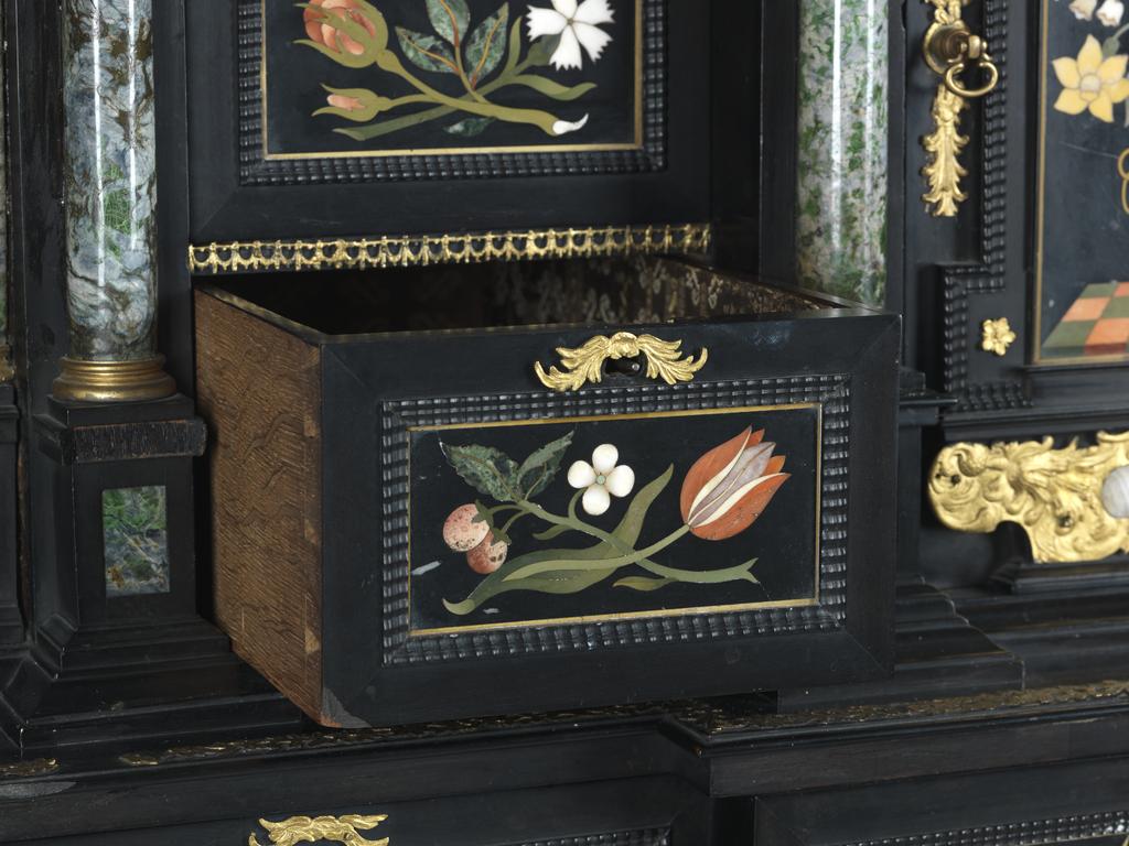 An image of Augsburg 17th century silver-gilt mounted ebony Table Cabinet with Florentine pietre dure plaques from the Grand Ducal workshops. Boscher, Elias (1600-1674). Of architectural form, the stepped superstructure fitted with three frieze drawers above a central cupboard door, architectural pediment and inset with a panel depicting a chaffinch on a fruiting bough, flanked by columns of Smaragdite Gabbro with Corinthian capitals flanked by two deep drawers, inset with panels depicting parrots within fruiting boughs, with three further fitted drawers below, the large central cupboard door inset with a panel of a lapis lazuli flower filled urn issuing stems including a carnation, anemone, daffodil and lilies and enclosing an architectural interior, flanked by further Smaragdite Gabbro Ionic columns and three drawers to each side inset with further symmetrical floral panels including tulips, roses, carnations, lilies and bluebells, on shaped bracket feet, each side inset with large central panels of parrots on fruiting boughs, with four further floral panels above and below, the reverse veneered in kingwood, amaranth and stained walnut in geometric stellar designs. The cabinet containing a complex series of fourty-four secret drawers and compartments to the interior. The cabinet is signed in pencil on the underside 'Elias Boscher' and 'gemacht'. The central cabochon mount stamped with the Augsburg town mark and the silversmiths mark of Johann Spitzmacher (active 1655-1678). The front bears the ebony stamp "EBEN" with the pinecone of Augsburg. Height 85 cm, width 84cm, depth 40cm, circa 1660.