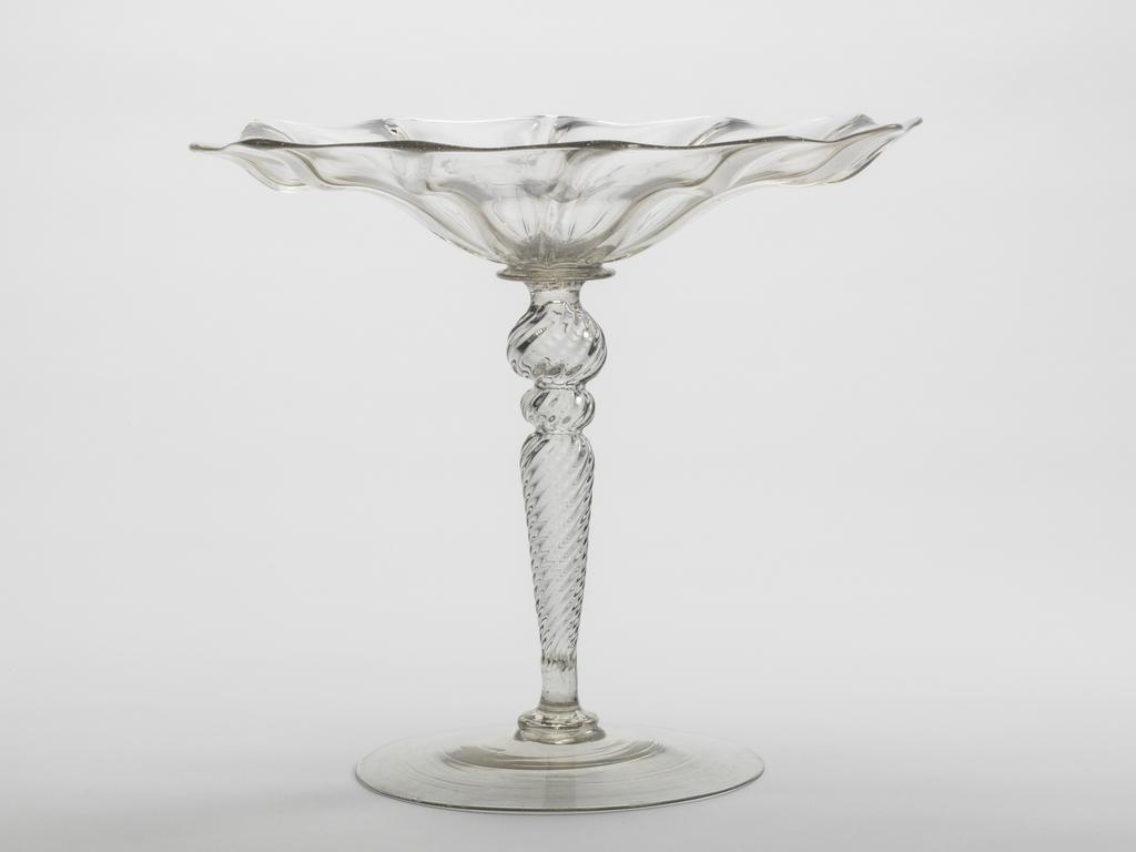 An image of Glass/Tazza. Unknown, glasshouse, The Veneto. Open flower like bowl with wavy edge, and ribbing on the underside. Inverted baluster stem with two knops, all ribbed spirally. Plain foot. Lead-glass, moulded, with ribbed decoration, height, whole, 15.7 cm, diameter, whole, 7.0 in, circa 1575-1625. Renaissance.