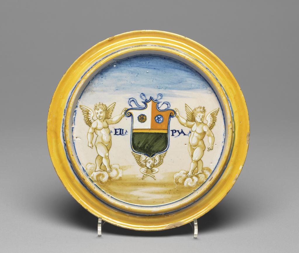 An image of The Holy Family. Maiolica cover from an accouchement set bowl, painted in polychrome with the Holy Family. Tagliere. Milan Marsyas Painter, possibly, The Marches, Urbino. Xanto, Francesco, painter, possibly, The Marches, Urbino. Caraglio, Giovanni Giacomo, printmaker, after. Parmigianino (Francesco Mazzola), painter, after, (Italian, 1503-1540). Circular with a flange on the underside; the upper surface flat with a convex rim. The Holy Family in an architectural setting; on the rim, bound leaves and berries between concentric yellow bands. On the back, two putti standing on clouds support a shield bearing the arms sable, a fess or, a chief party per pale gules and argent two rosettes counterchanged flanked by the letters `ELI' and `PYA'; below, a winged putto's head. Pale buff earthenware, tin-glazed overall. Painted in blue, turquoise-green, yellow, orange, black, grey, and white, height, whole, 1.9 cm, diameter, whole, 19.2 cm, circa 1531. Renaissance. Production Note: If not painted by the 'Milan Marsyas Painter' this was probably painted by Francesco Xanto Avelli da Rovigo. The Holy Family on the top of the cover was derived from the Adoration of the Shepherds, engraved by G.G. Caraglio after Parmigianino.