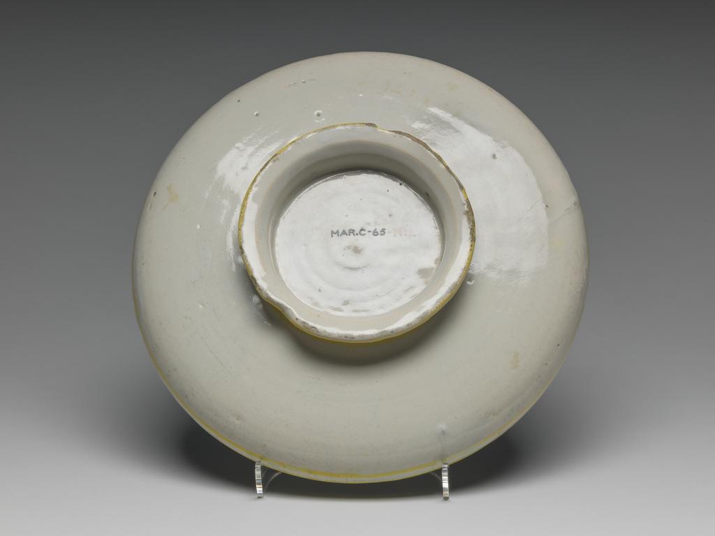 An image of Maiolica. Shallow bowl on low foot. Unidentified potter. This type of dish is traditionally attributed to Castel Durante, but could have been made in Urbino. A bust of a young woman, three-quarters to right, her gaze cast downwards to her right. Her blonde hair is dressed in a plait over the crown of her head. She wears a green and yellow turban embroidered with black scrolling foliage; two gold chains and a rope of beads; and a green dress with a yellow chemise showing on the shoulders. A scroll inscribed `LAVRA/BELLA' in blue projects on either side of her head. The background is blue. On the back are four concentric yellow bands: on the edge of the foot, just beyond the junction of the foot and bowl, and two round the rim. Pale buff earthenware, tin-glazed overall. Painted in blue, green, yellow, orange, cream, and black. Height, whole, 4.7 cm, diameter, whole, 22.7 cm, circa 1525-1535. Renaissance.