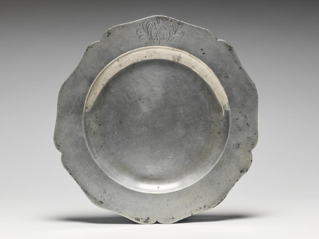 An image of Plate. Thomas Chamberlain of King Street, Soho. Pewter, cast and hammered, with engraved arms, height, whole, 2 cm, diameter, whole, 25 cm, circa 1750-1775. Circular with five indentations and five outward curves, curved sides and flat base. The underside of the rim has an applied raised edge. The rim is engraved with tan almost circular shield bearing he unregistered coat-of-arms of David Garrick (1717-79), surrounded and foliage. This plate was part of a service made for the celebrated actor and manager of the Drury Lane Theatre, David Garrick (1717-79). If the arms had tinctures (colours) they would be described: 'Per pale or and azure on the dexter compartment a tower gules and on the sinister on a mount vert a sea horse azure mane, fins and tail of the first, on a chief gold, three mullets of the second. (See Documentation, Burke) Thirty-one pieces from the service bearing either Garrick's arms or crest are at the Shakespeare Birthplace Trust at Stratford-on-Avon. A.F. de Navarro Bequest.