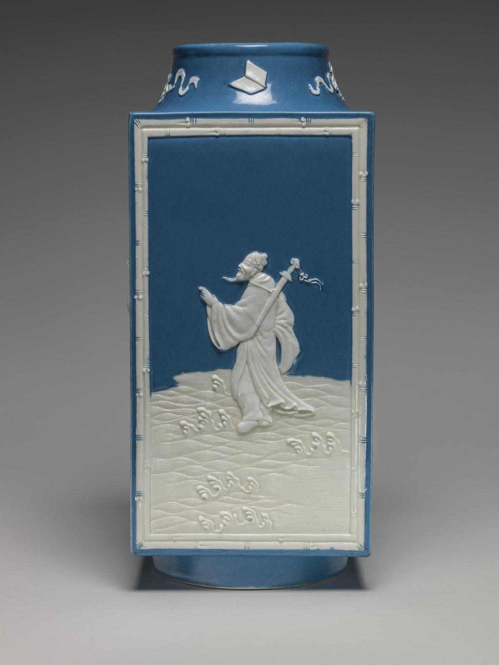 An image of Vase. One of two square stoneware vases, with a sloping cylindrical neck and cylindrical foot ring. With applied white reliefs and opaque powder blue glaze on the interior and parts of the exterior. Decorated on the sides with four of the Eight Immortals standing on waves, enclosed by a rectangular bamboo border, and on the neck with four symbols, all in white. The immortals are Lu Dong-bin (with sword), Li Tie-guai (with crutch and gourd), Lan Cai-he (with basket of flowers) and Zhang Guo-lao (with rods in cane). Mark: Six character seal script mark of Qianlong. Stoneware, powder blue glaze, with applied relief, height 28.5 cm, width 12.5 cm. Qianlong Period (1736-1795). Chinese.