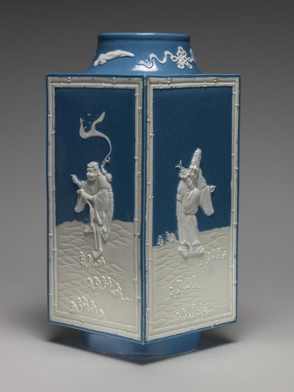 An image of Vase. One of two square stoneware vases, with a sloping cylindrical neck and cylindrical foot ring. With applied white reliefs and opaque powder blue glaze on the interior and parts of the exterior. Decorated on the sides with four of the Eight Immortals standing on waves, enclosed by a rectangular bamboo border, and on the neck with four symbols, all in white. The immortals are Lu Dong-bin (with sword), Li Tie-guai (with crutch and gourd), Lan Cai-he (with basket of flowers) and Zhang Guo-lao (with rods in cane). Mark: Six character seal script mark of Qianlong. Stoneware, powder blue glaze, with applied relief, height 28.5 cm, width 12.5 cm. Qianlong Period (1736-1795). Chinese.