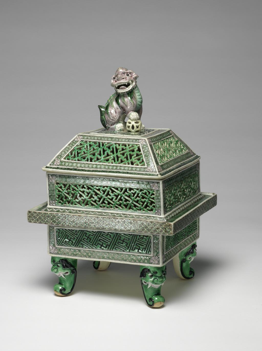 An image of Perfume burner and cover/cassolette. Famille verte, biscuit. The two-storied body is of square section with upright sides supported on four green scroll legs issuing from ferocious animal masks. The lower part of the body has raised rectanglar panels incised with a wan-diaper pattern picked out in black on a green ground, reserved against an intricate green and yellow trellis diaper, surmounted by a galleried flange with a florette diaper in green, yellow and aubergine round the edge and a green-ground spiral scroll on the top. The upper part of the body is intricately pierced with green wan-diaper panels reserved against pale green cell-diaper. There is a matching pierced wan-diaper and cell-diaper pattern on the sloping sides of the domed cover which has a flat top, centred with a large pale aubergine Buddhist lion knop, the animal's right paw resting on a pale yellow brocade ball. The interior and the base are unglazed. Hard-paste porcelain, pierced and applied, and painted on the biscuit in an enamel palette of black, yellow, aubergine and two tones of green, height, whole, 29.5 cm, diameter, whole, 19.6 cm, circa 1662-circa 1722. Late 19th century. Qing Dynasty (1644-1912). Chinese.
