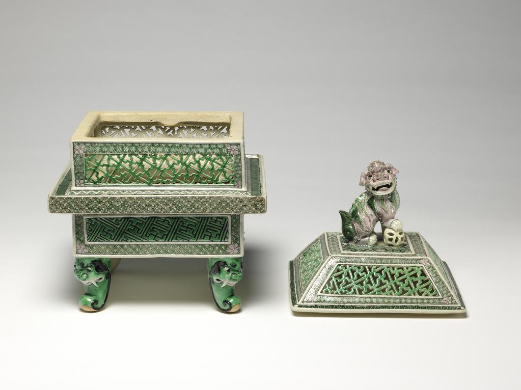 An image of Perfume burner and cover/cassolette. Famille verte, biscuit. The two-storied body is of square section with upright sides supported on four green scroll legs issuing from ferocious animal masks. The lower part of the body has raised rectanglar panels incised with a wan-diaper pattern picked out in black on a green ground, reserved against an intricate green and yellow trellis diaper, surmounted by a galleried flange with a florette diaper in green, yellow and aubergine round the edge and a green-ground spiral scroll on the top. The upper part of the body is intricately pierced with green wan-diaper panels reserved against pale green cell-diaper. There is a matching pierced wan-diaper and cell-diaper pattern on the sloping sides of the domed cover which has a flat top, centred with a large pale aubergine Buddhist lion knop, the animal's right paw resting on a pale yellow brocade ball. The interior and the base are unglazed. Hard-paste porcelain, pierced and applied, and painted on the biscuit in an enamel palette of black, yellow, aubergine and two tones of green, height, whole, 29.5 cm, diameter, whole, 19.6 cm, circa 1662-circa 1722. Late 19th century. Qing Dynasty (1644-1912). Chinese.