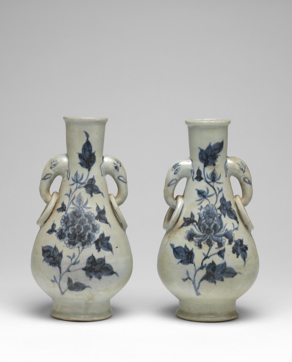 An image of Vase with elephant handles. Unknown maker, China, Folk kilns. The pear-shaped vase has a slender neck, thick, rounded lip, and two elephant head and fixed-ring handles. The body tapers to a deep, flared foot with deep flat, recessed base. The elephant handles are finely painted with eyes, eyelashes, tusks and ears, and the fixed rings with single circle ornament. On each side of the body are elegant peony sprays in blue. The interior is unglazed, as is the base which is covered with a red, sandy deposit. Hard-paste porcelain, thrown, painted in underglaze blue, with base and interior unglazed. Height, whole, 24.6 cm, diameter, whole, 12.7 cm, circa 1300-1368. Yuan Dynasty (1206-1368). Collection: H.S. Reitlinger. Of a pair of vases, with C.689B-1991.