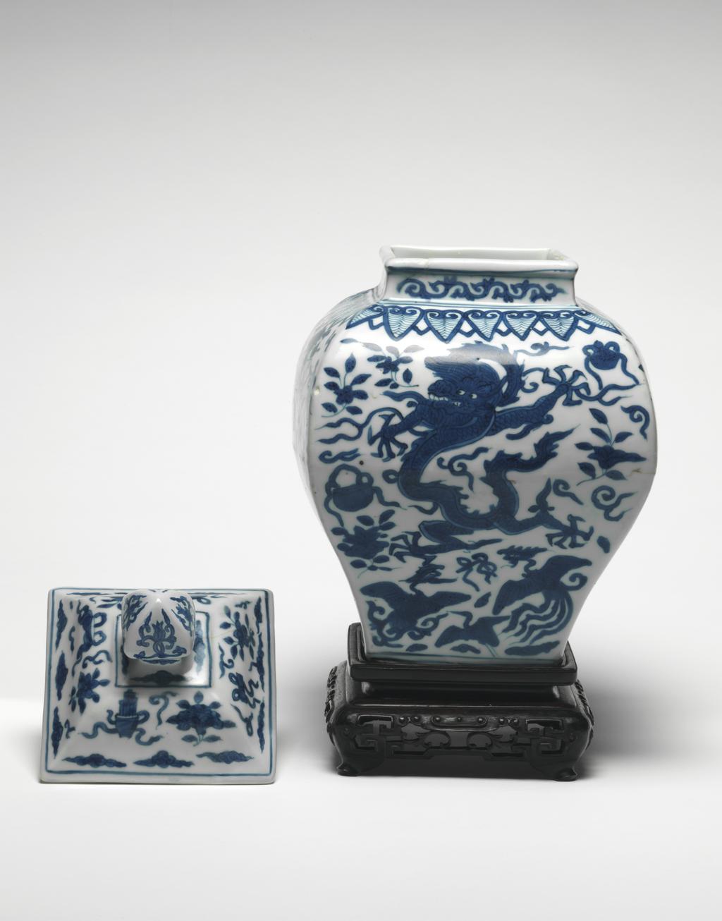 An image of Square section baluster jar and cover with wooden stand. The jar is painted on all sides with five-claw dragons, phoenixes and the eight precious emblems below a border of ruyi head lappets around the shoulders and scrolls on the neck. The cover is painted with the eight precious emblems and cloud scrolls. Hard-paste porcelain, thrown, decorated in underglaze-blue, height, jar, 26.6 cm, height, whole, 32.0 cm, diameter, whole, 17.6 cm, circa 1573-1619. Ming Dynasty (1368-1644), Wanli period (1573-1619). Chinese. As a pair with C.716.2 & A & B-1991.