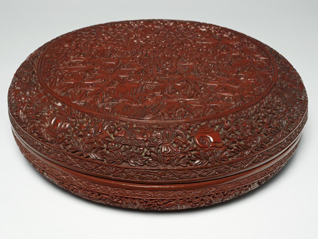 An image of Wooden box, lacquered in red on an ochre ground, with a black interior. The exterior is carved with a praying mantis and other insects, as well as a lizard and a toad. Wood, lacquering, carving, height, 8.8 cm, diameter, 38.8 cm, possibly early 16th century. Ming Dynasty. Chinese.