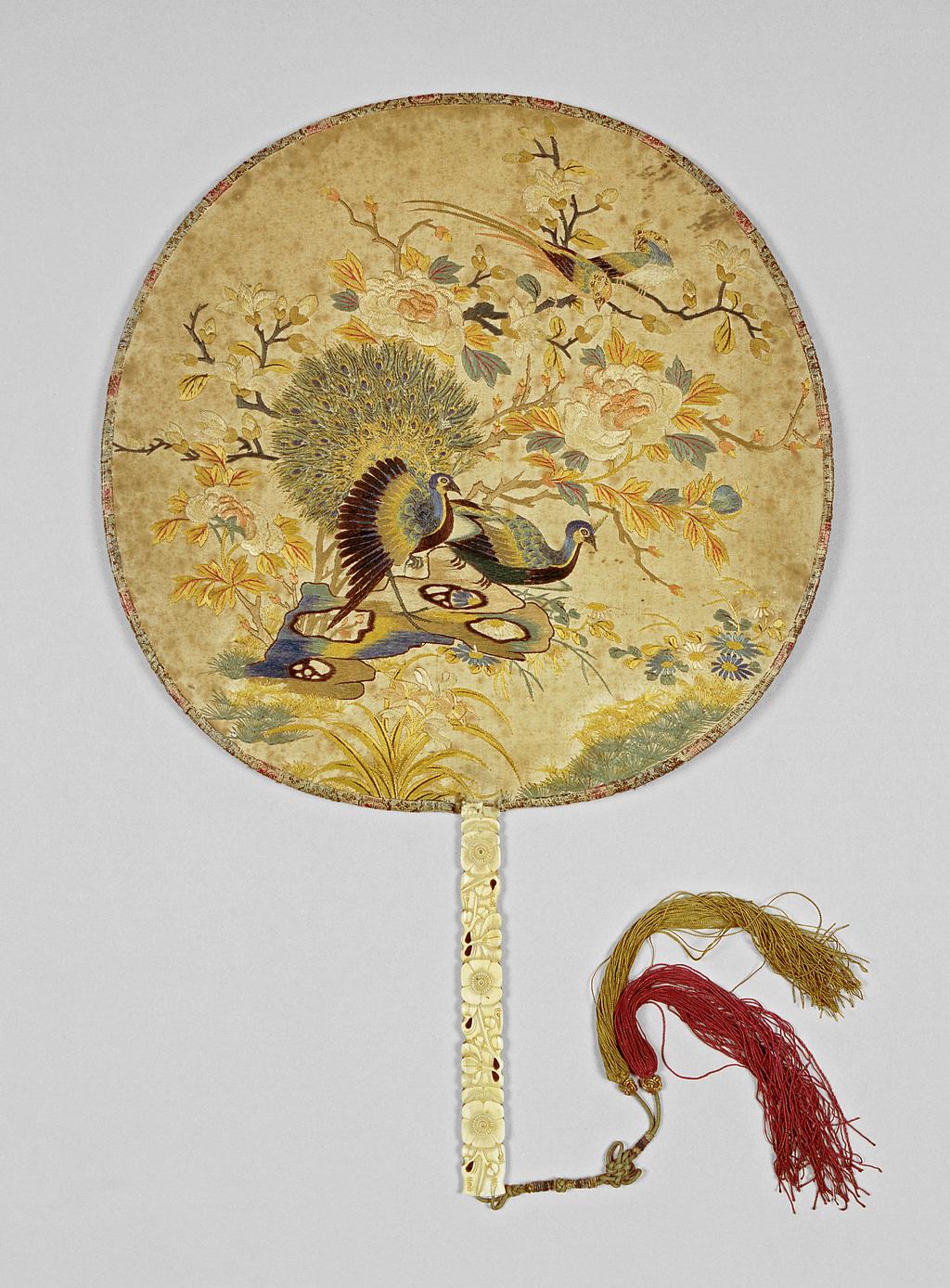 An image of Screen fan/pien mien. Unknown maker, China. Decorated with peacocks and orioles with flowering tree peonies, magnolias and rocks, the stick formed as flowering prunus. Stick: ivory carved in relief and pierced; knotted string of green silk bound in blue, with red and gold tassels, with sliding gilt paper-wrapped toggles. Rim: brocade covered. Face: polychrome silk embroidered on satin. Height, guards, 40.3 cm, width, whole, 26.6 cm, after 1770 to before 1830. Collection: Messel-Rosse Collection.