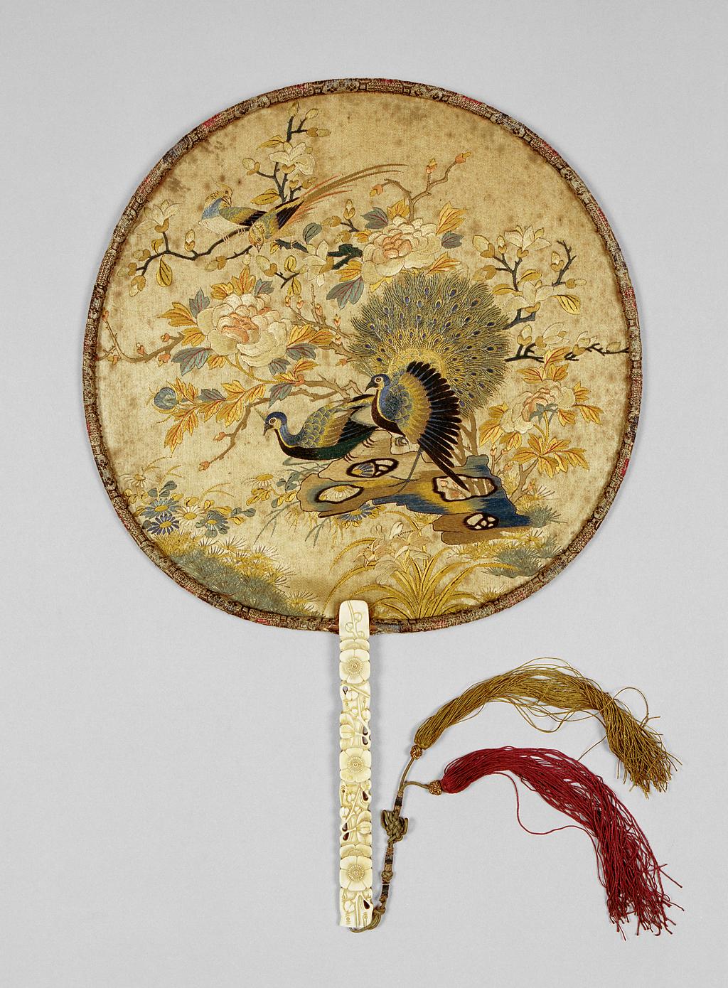 An image of Screen fan/pien mien. Unknown maker, China. Decorated with peacocks and orioles with flowering tree peonies, magnolias and rocks, the stick formed as flowering prunus. Stick: ivory carved in relief and pierced; knotted string of green silk bound in blue, with red and gold tassels, with sliding gilt paper-wrapped toggles. Rim: brocade covered. Face: polychrome silk embroidered on satin. Height, guards, 40.3 cm, width, whole, 26.6 cm, after 1770 to before 1830. Collection: Messel-Rosse Collection.
