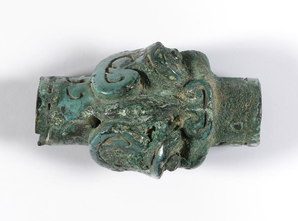 An image of Finial. Unknown maker, China. In the form of a human head with a flowing headdress, on which is a conventionalised bird. Covered with a strong green patina. Bronze, height 7 cm, length 7 cm, 1000 to 900 B.C. Chou dynasty (1122-249 BC).