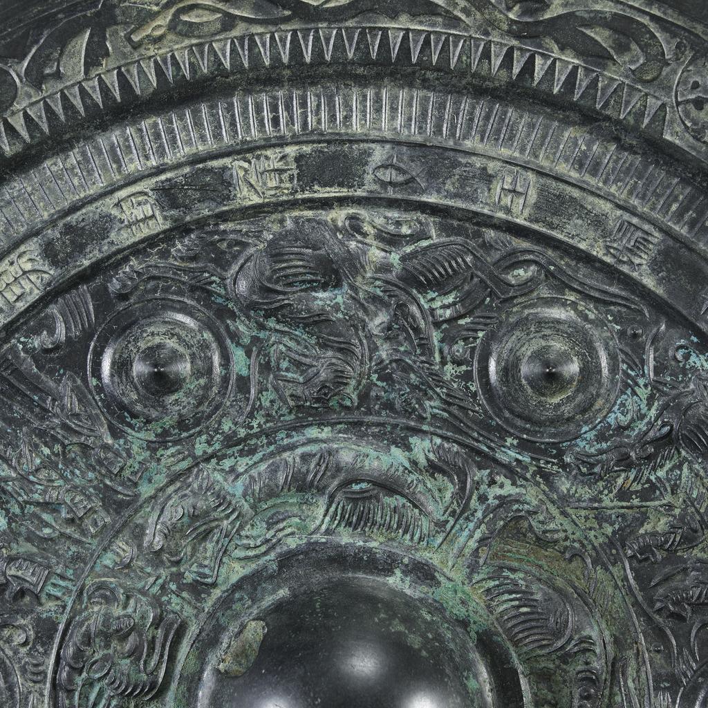 An image of Relief. Mirror made from bronze, inscribed with makers name (Teh'e). Around the central boss is a band of tail-less dragons, surmounted by a band of dragons, figures of men, and chariots. There is a band of inscription and a rim with dragons in light relief. Diameter 21.6 cm. Late Han Dynasty (206 BC-220 AD).