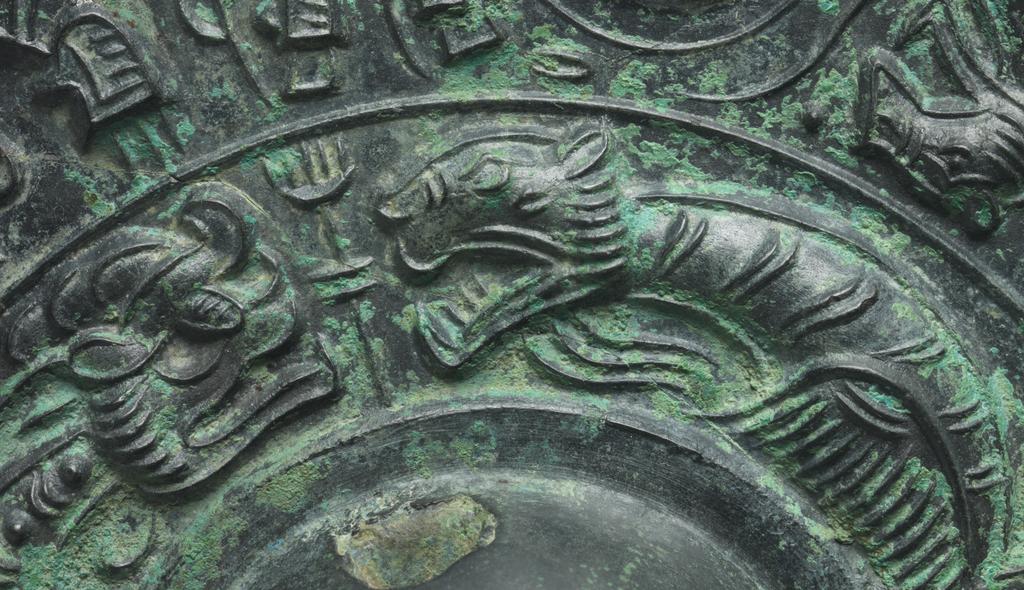 An image of Relief. Mirror made from bronze, inscribed with makers name (Teh'e). Around the central boss is a band of tail-less dragons, surmounted by a band of dragons, figures of men, and chariots. There is a band of inscription and a rim with dragons in light relief. Diameter 21.6 cm. Late Han Dynasty (206 BC-220 AD).