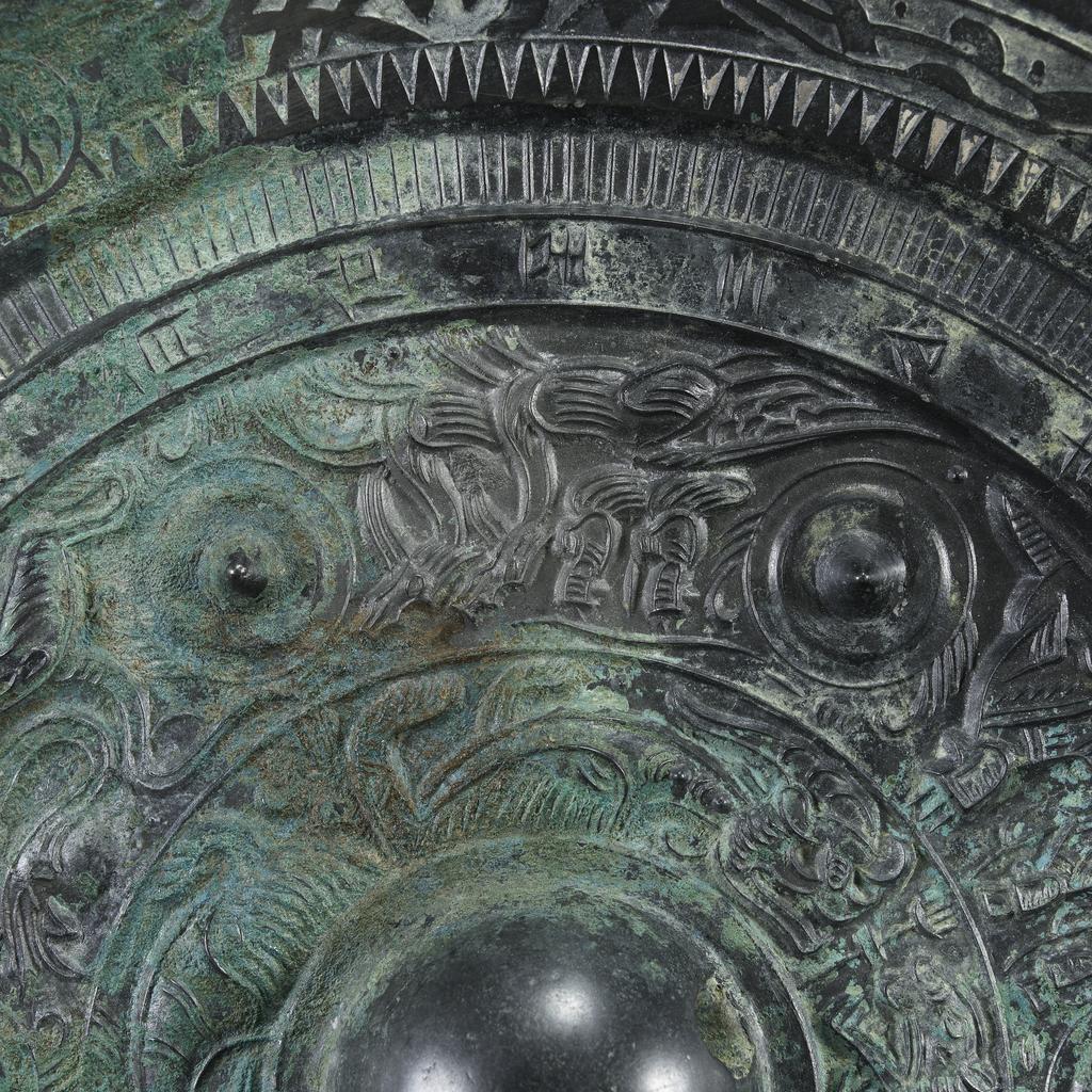 An image of Relief. Mirror made from bronze, inscribed with makers name (Teh'e). Around the central boss is a band of tail-less dragons, surmounted by a band of dragons, figures of men, and chariots. There is a band of inscription and a rim with dragons in light relief. Diameter 21.6 cm. Late Han Dynasty (206 BC-220 AD).