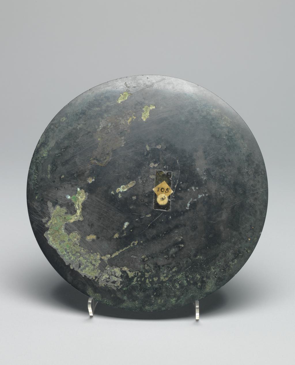 An image of Relief. Mirror made from bronze, inscribed with makers name (Teh'e). Around the central boss is a band of tail-less dragons, surmounted by a band of dragons, figures of men, and chariots. There is a band of inscription and a rim with dragons in light relief. Diameter 21.6 cm. Late Han Dynasty (206 BC-220 AD).