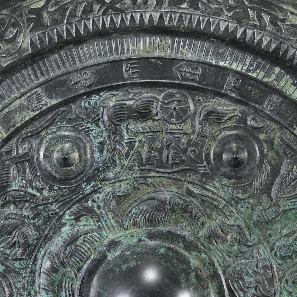 An image of Relief. Mirror made from bronze, inscribed with makers name (Teh'e). Around the central boss is a band of tail-less dragons, surmounted by a band of dragons, figures of men, and chariots. There is a band of inscription and a rim with dragons in light relief. Diameter 21.6 cm. Late Han Dynasty (206 BC-220 AD).