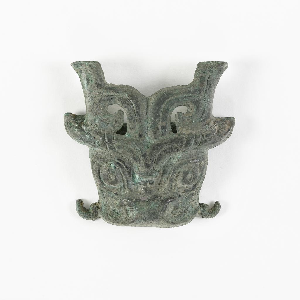An image of Unknown maker, China. T'ao t'ieh mask as a buckle, with a cross-piece at the back for attachment. The mask has horns and ears, with whiskers at the side of the mouth. It has a rough, grey-green patina. Bronze, width 5.4 cm, 700- 600 B.C. Chou dynasty (1122-249 BC).