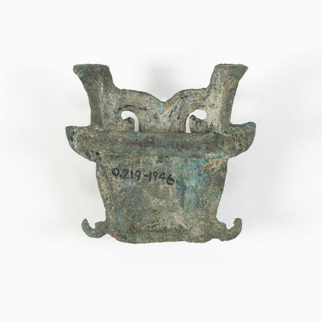 An image of Unknown maker, China. T'ao t'ieh mask as a buckle, with a cross-piece at the back for attachment. The mask has horns and ears, with whiskers at the side of the mouth. It has a rough, grey-green patina. Bronze, width 5.4 cm, 700- 600 B.C. Chou dynasty (1122-249 BC).