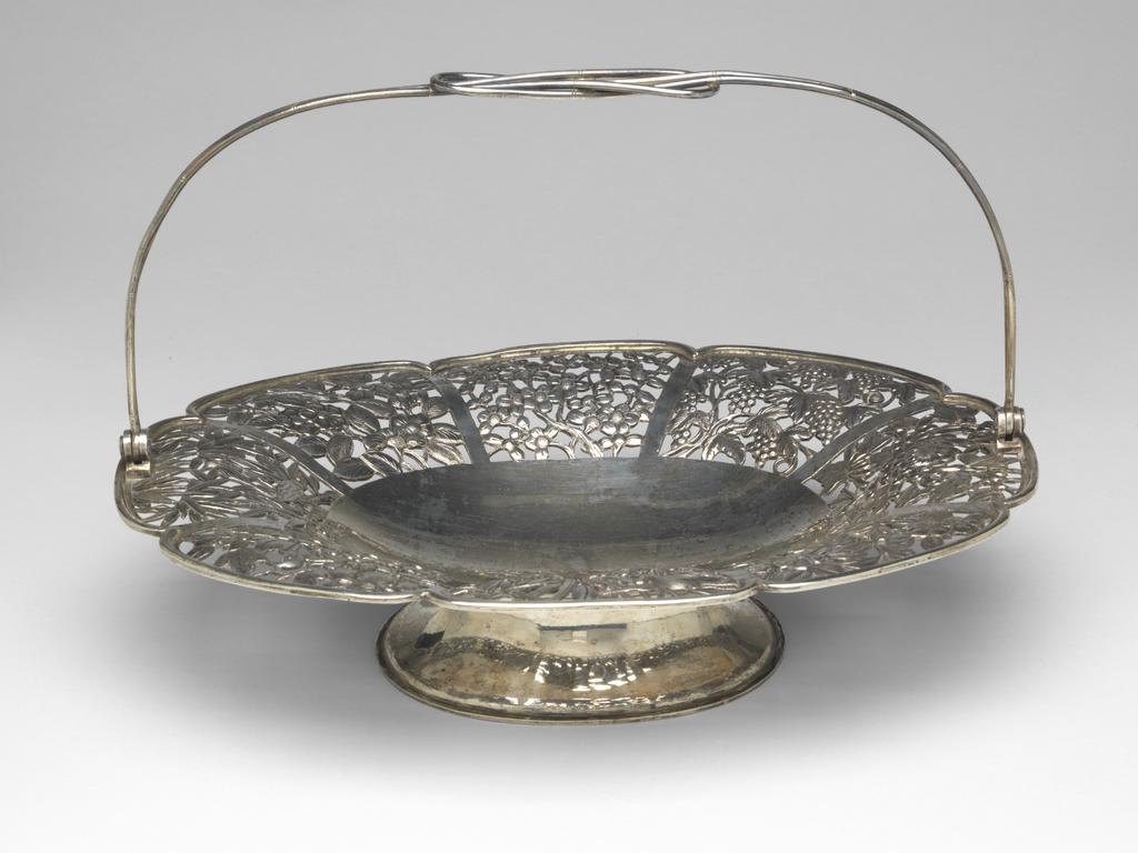 An image of Cake basket. Kwong Man Shing, maker, China, Hong Kong. The sides of the basket are pierced in eight panels of flowers (chrysanthemum, prun flowers, iris, bamboo and camellia) and separated by chased bands. The hinged central swing handle tapered at either end, is designed as bamboo stick. In the middle of the base impressed with ‘KMS (Kwong Man Shing, Hong Kong)’. Silver, the base and flared sides of the oblong basket are raised from a single sheet and seamed to a flared foot rim. Circa 1900.