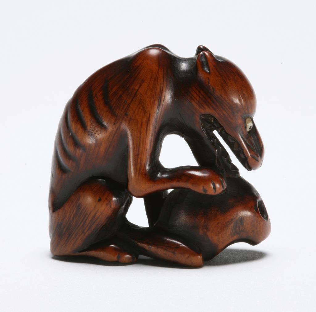 An image of Netsuke. A bony starving wolf seated gnawing at a skull. Unknown maker, Japan. Boxwood,  the eyes of the wolf are inlaid with ivory, 1770-1800.
