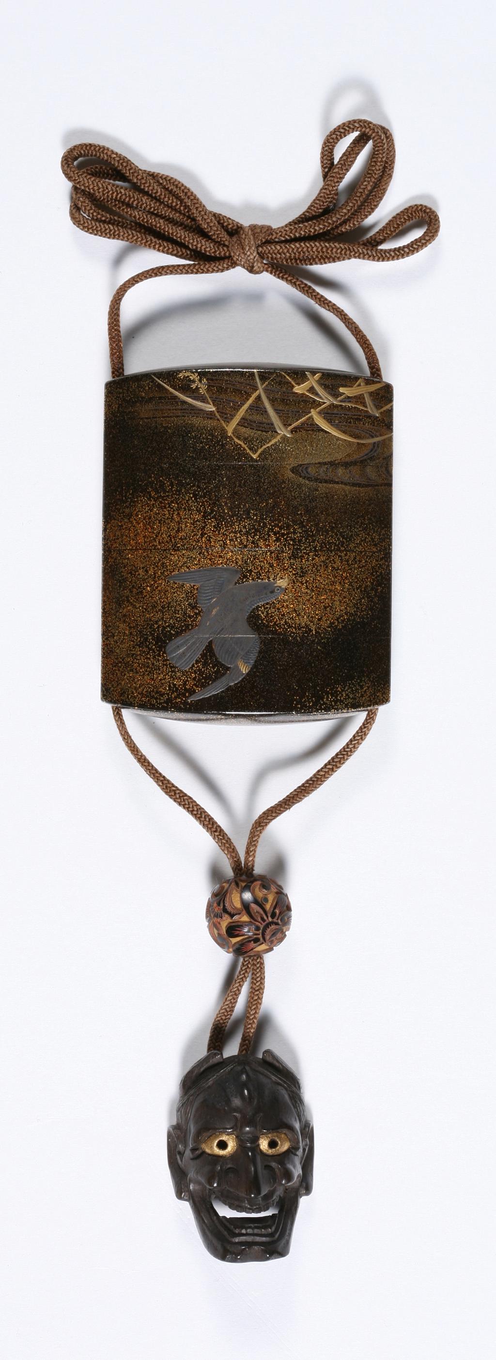 An image of Inro. Ojime of mask and netsuke. Unknown maker, Japan. Lacquer inro with four divisions, and with ojime and netsuke. Black ground with nashiji, decorated in gold and silver/pewter. Depicting an eagle diving towards a crane taking cover in long grasses. Risers and lining nashiji.  Ojime made from carved wood, lacquered in red and black. Mask netsuke of wood, with gold eyes. Height 7 cm, width 5.7 cm, 1700-1868. Edo Period (1615-1868). 